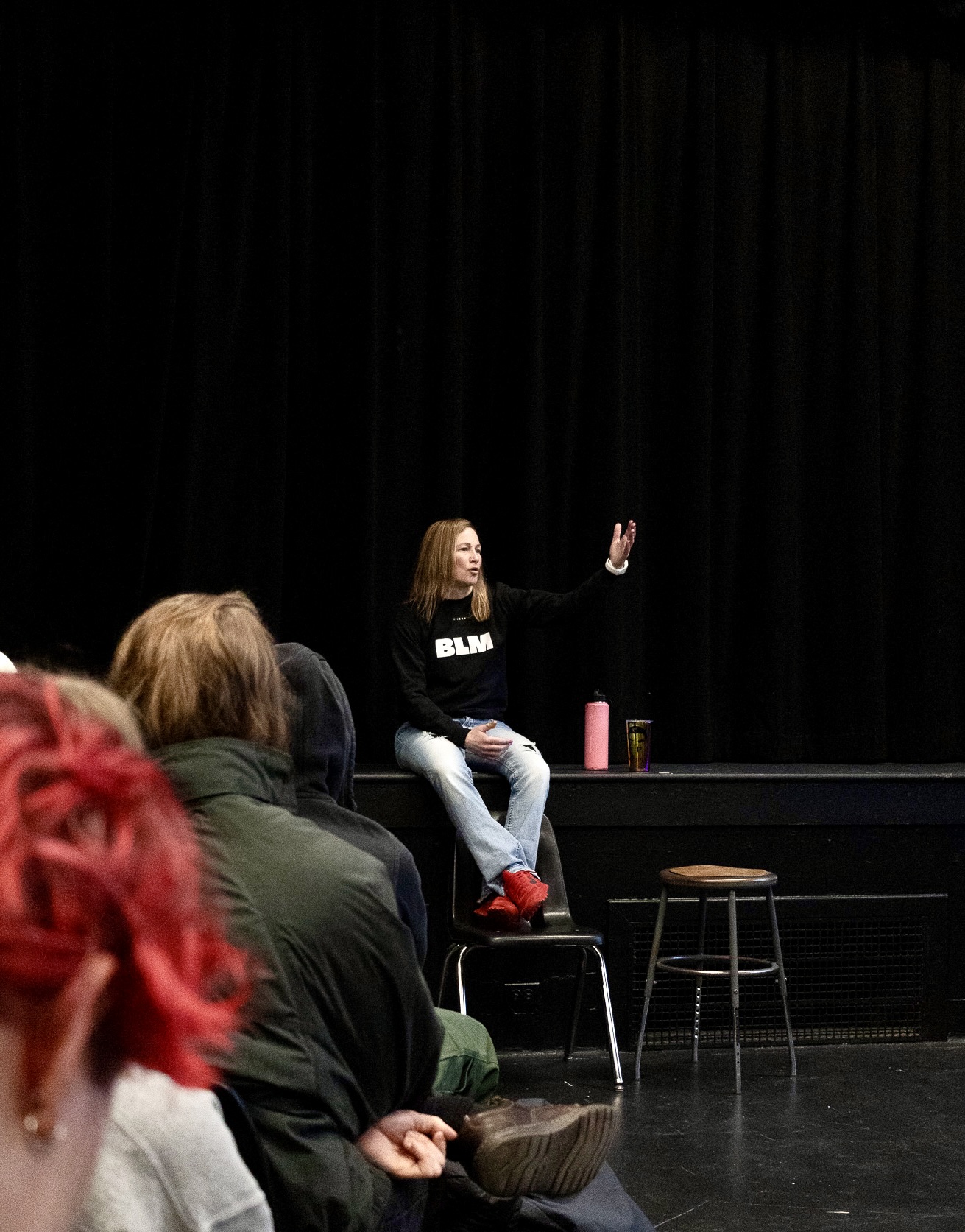 Person sitting on a stool on stage, raising hand, with audience in foreground, black curtain background.