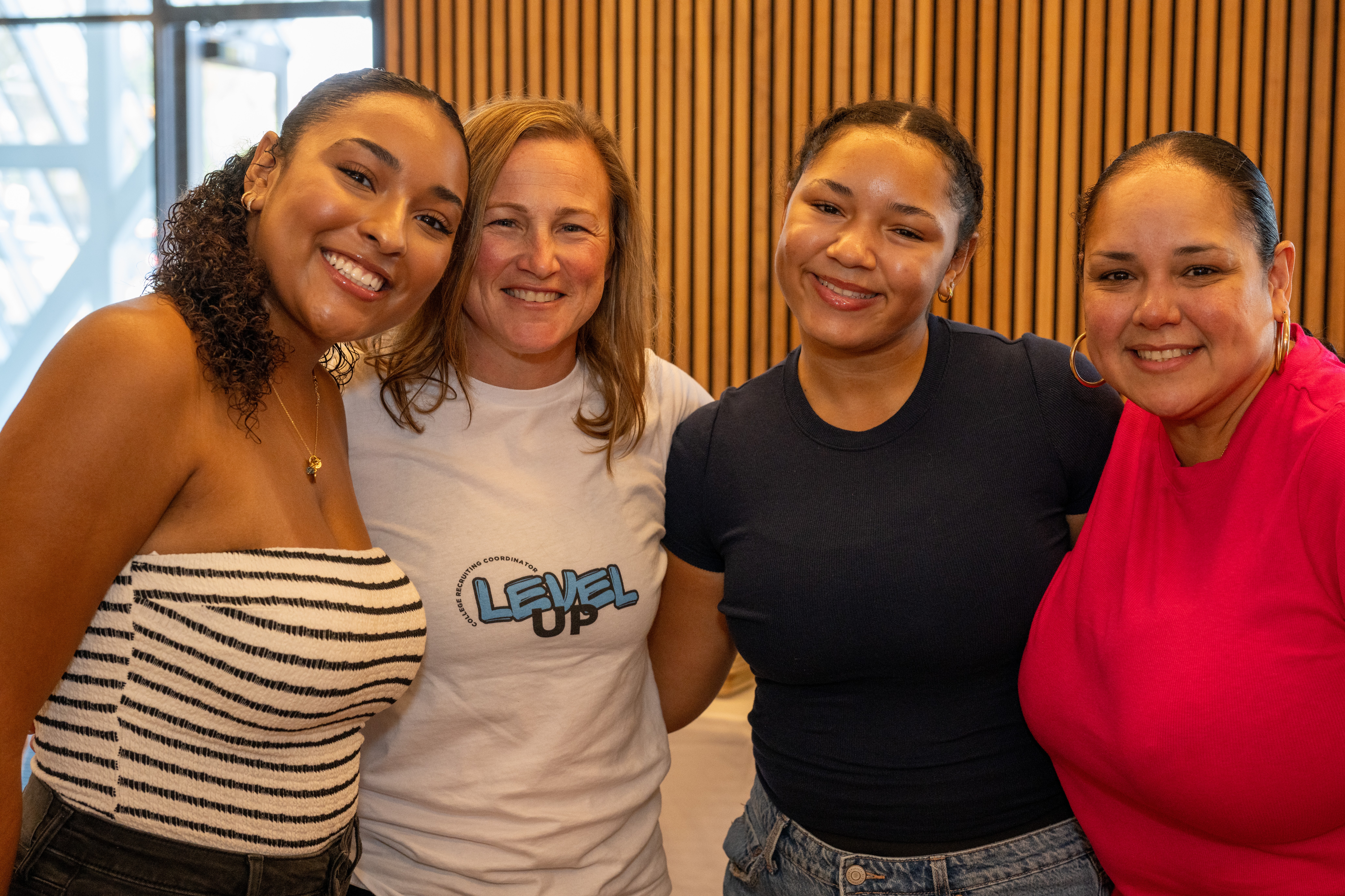 Four women smiling together indoors, standing close, with a wooden wall background.