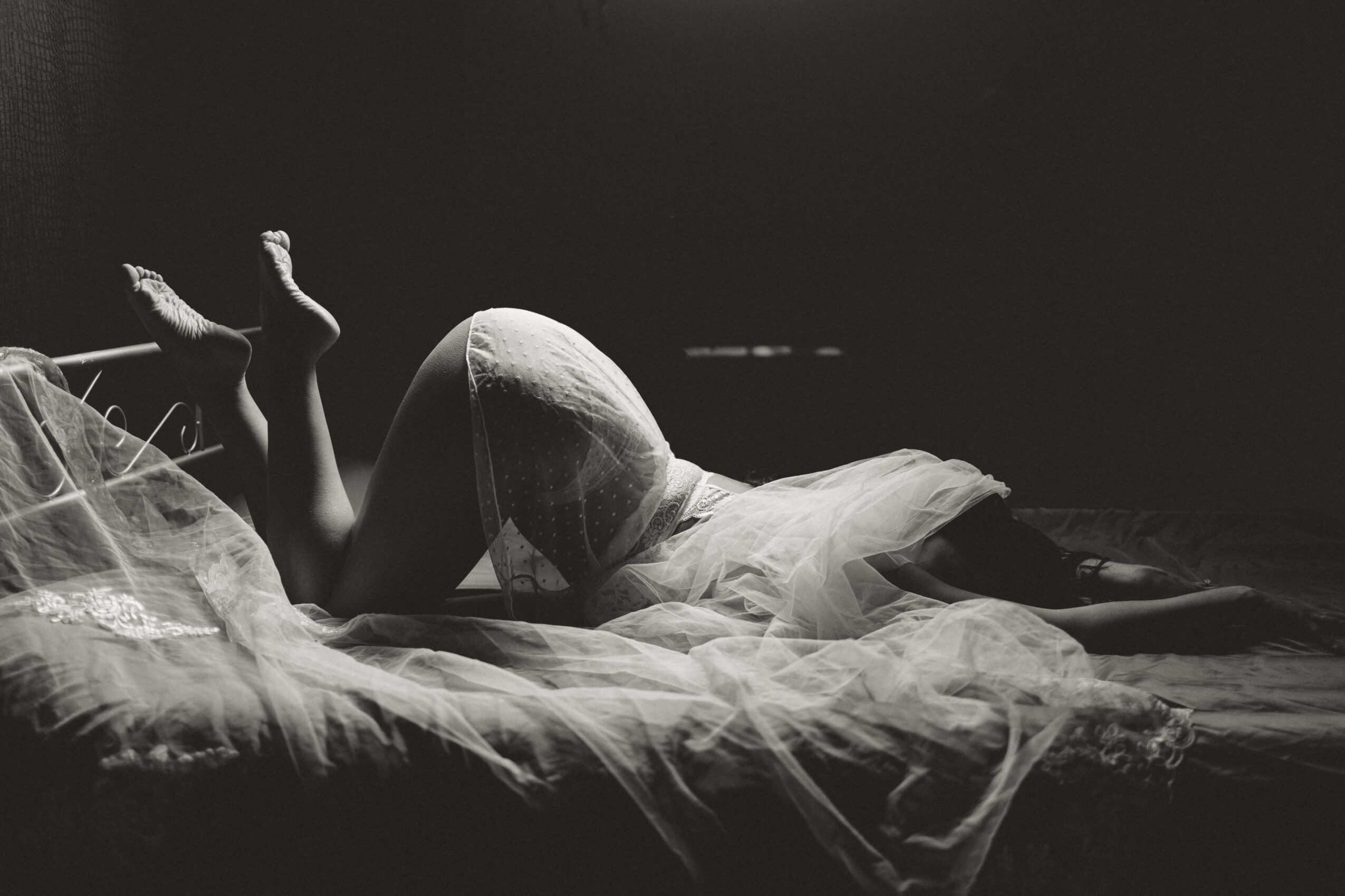 Person lying on bed with legs raised, covered by a sheer fabric, in a dark room, black and white photo.