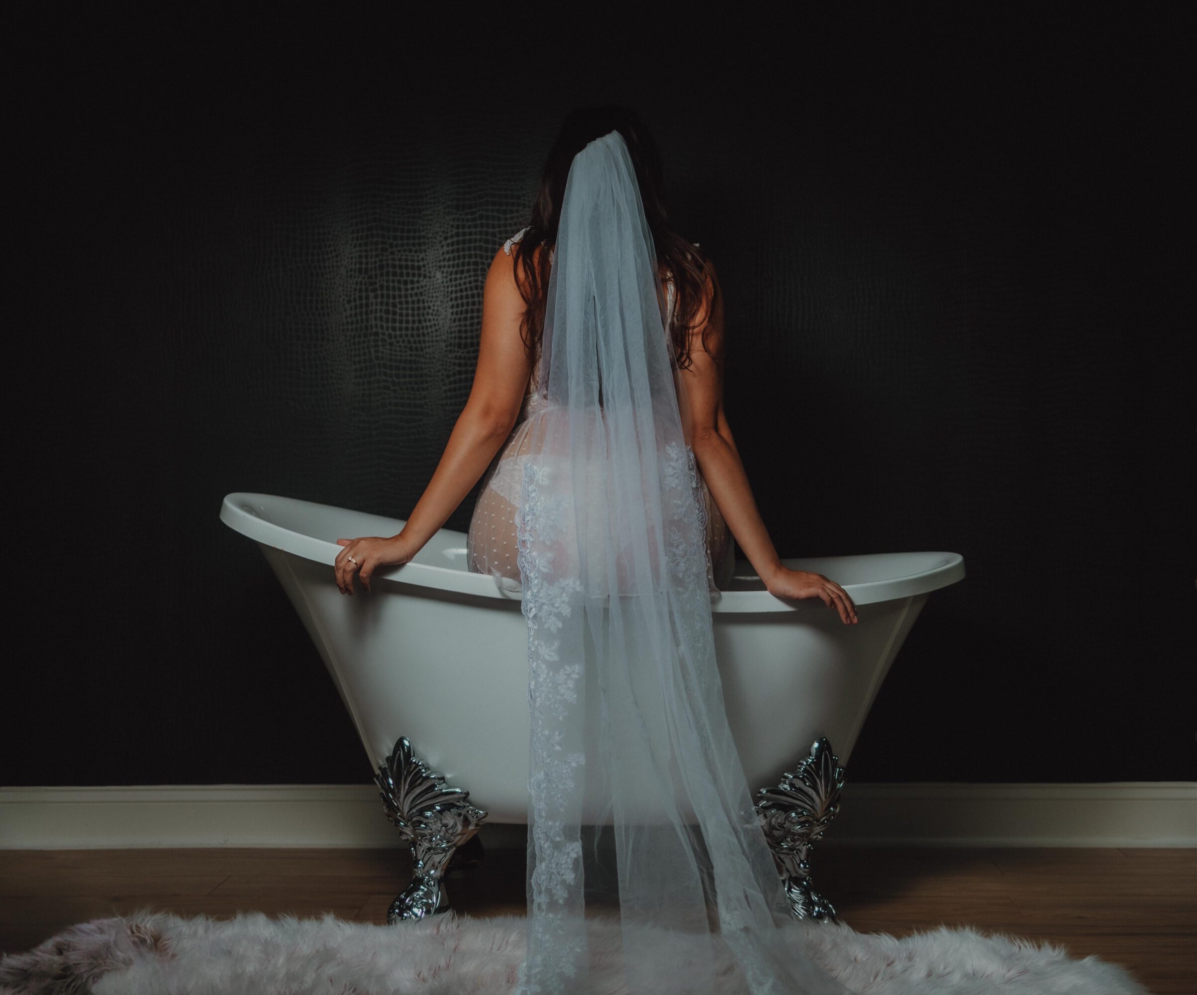 Person with a veil standing in a bathtub, facing away, against a dark wall.