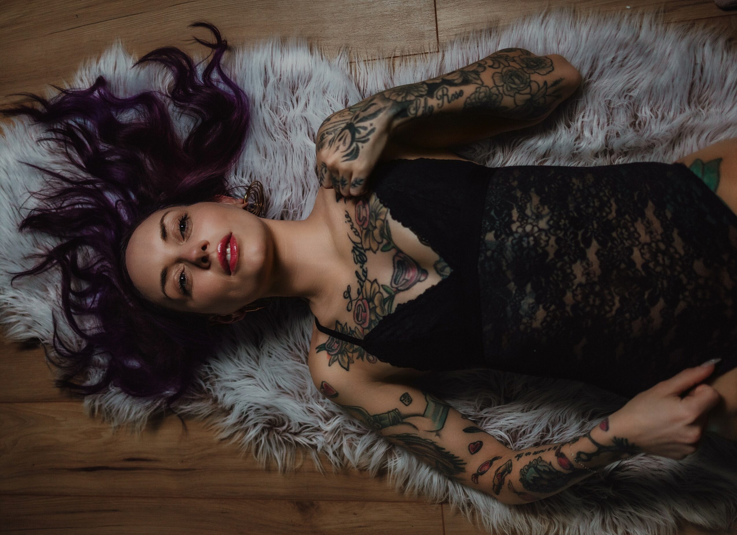 Woman with purple hair lying on a fur rug, wearing a black lace dress, with tattoos on her arms and chest.