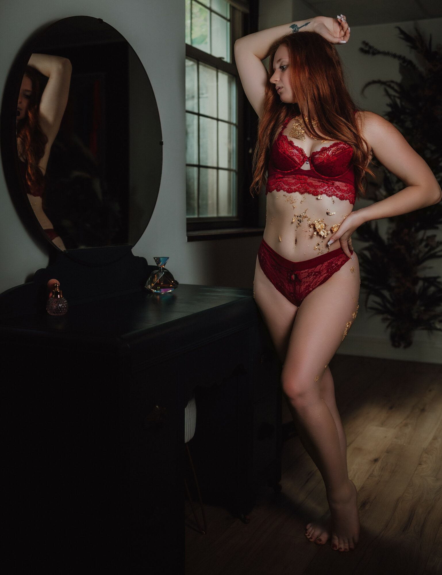 Woman in red lingerie standing near a window and mirror, with her hand on her head, in a room with dark furniture.