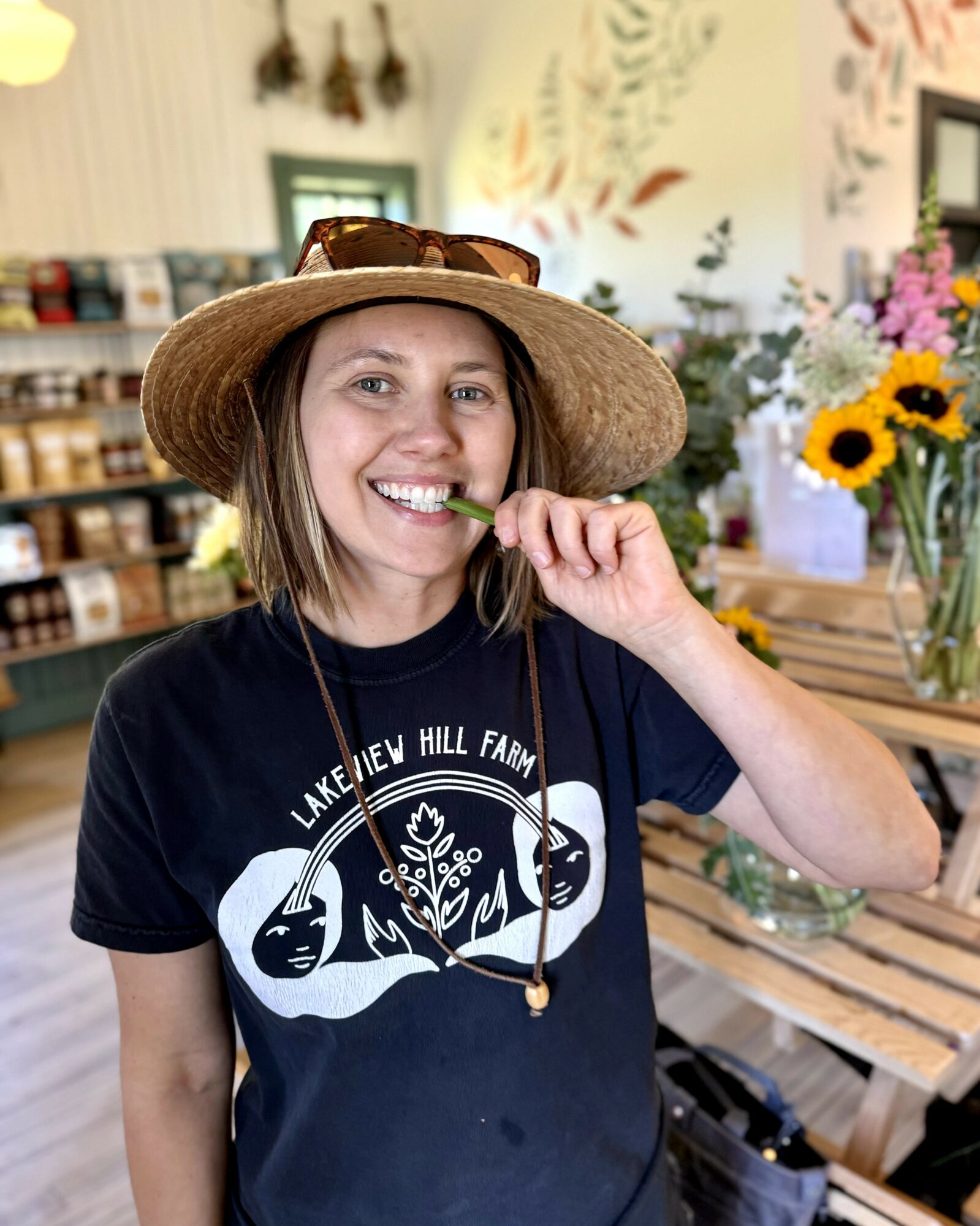 Woman wearing a wide-brimmed hat and black T-shirt, smiling and brushing teeth in a store with flowers and books.