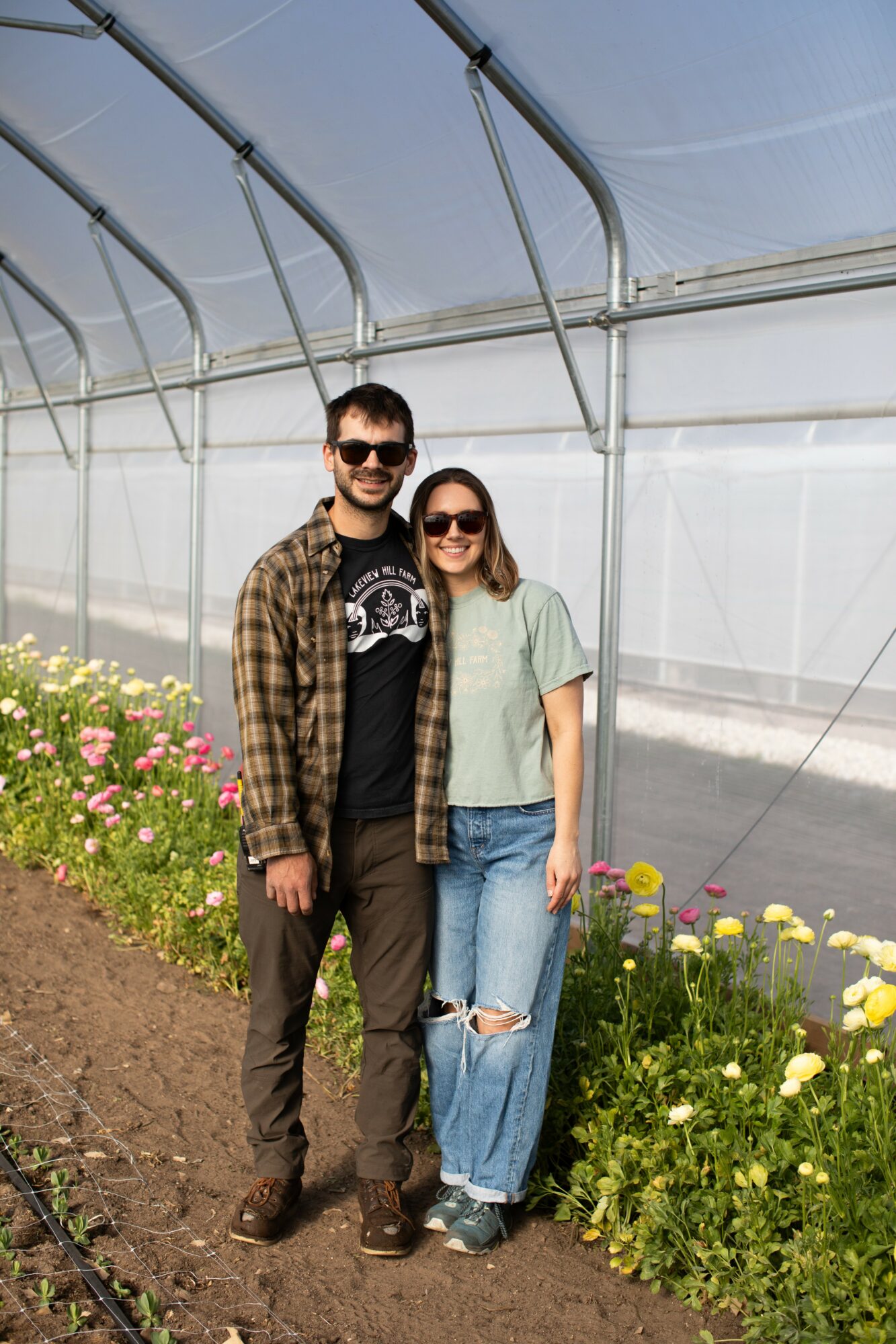 A man and woman stand inside a greenhouse with flowers along the path, smiling at the camera.