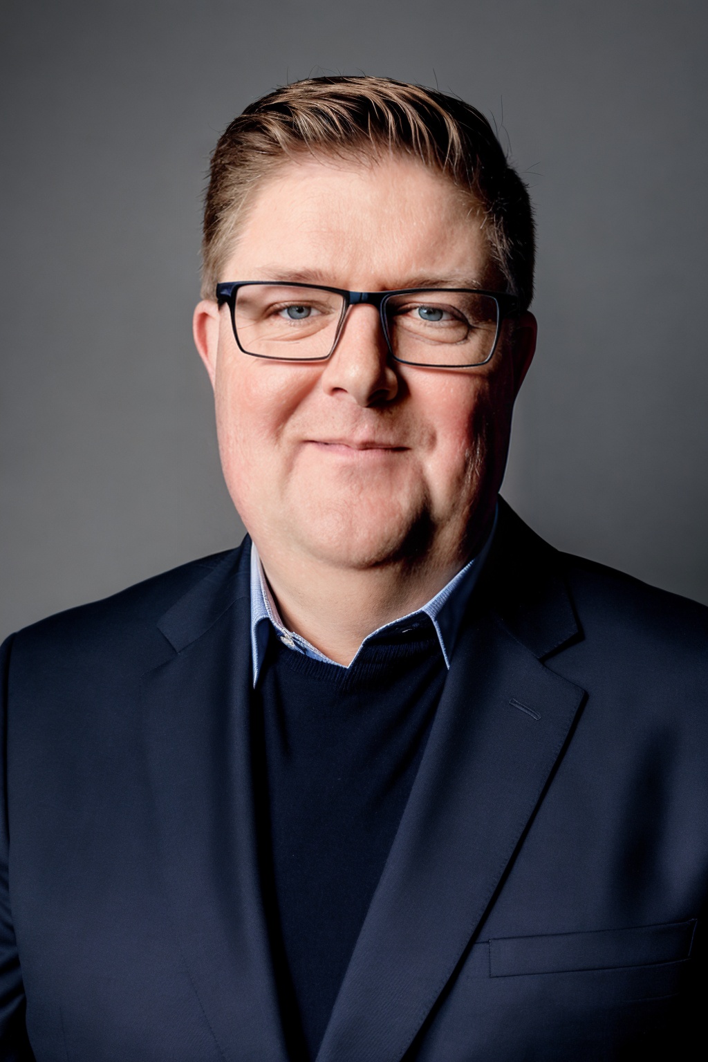 Portrait of a man with glasses, wearing a dark suit and collared shirt, against a gray background.