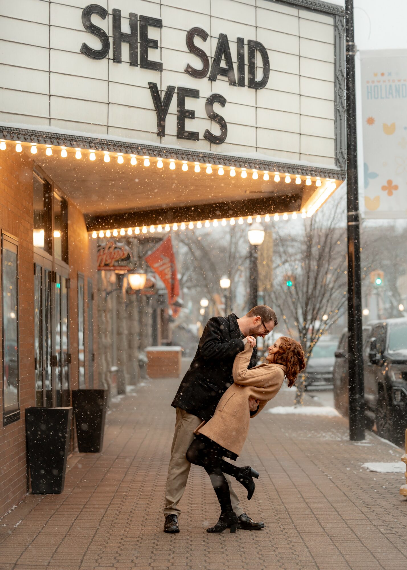 A couple dancing on a city sidewalk in front of a building with a sign that reads 'SHE SAID YES'.