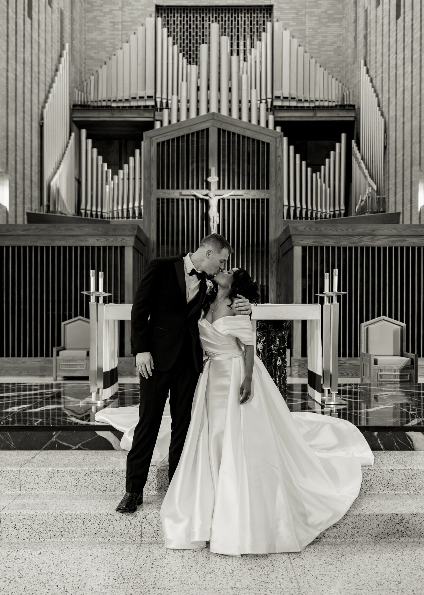 A bride and groom stand together in front of an altar with a large pipe organ in the background, holding hands.