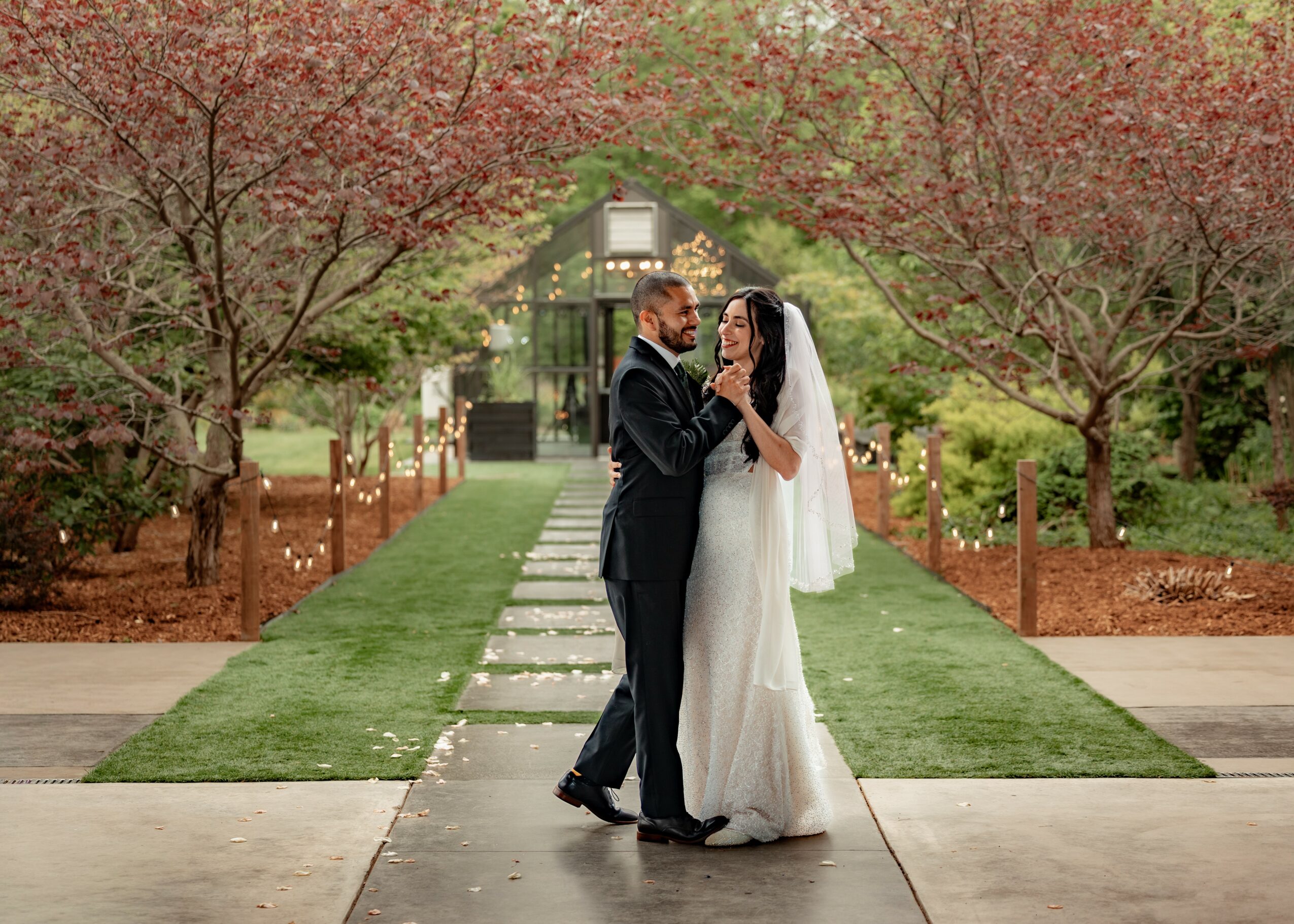 A bride and groom stand close together on a garden pathway, surrounded by trees with red and green leaves, during a wedding.