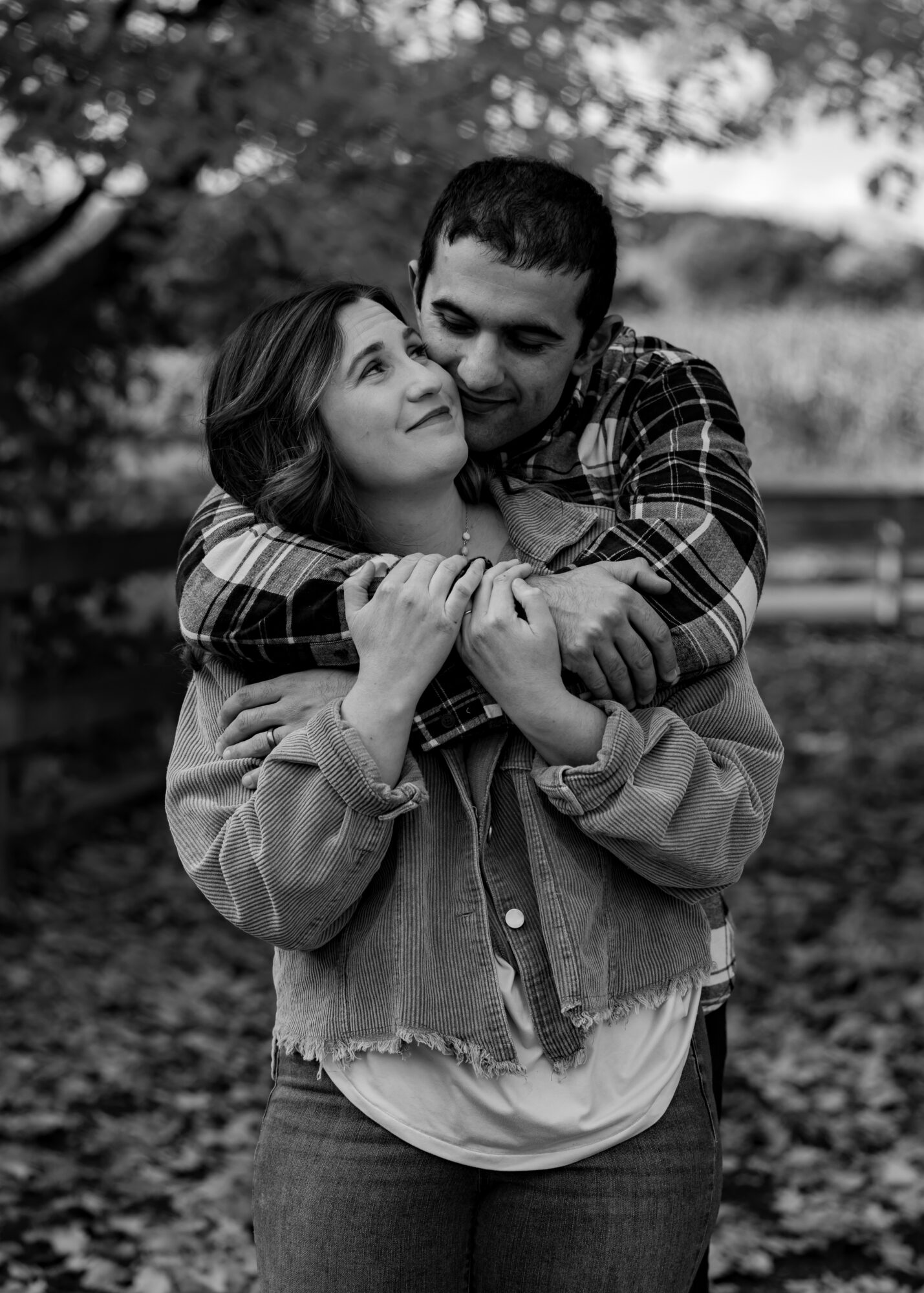 A couple embraces outdoors, smiling and looking at each other, with trees and a fence in the background.