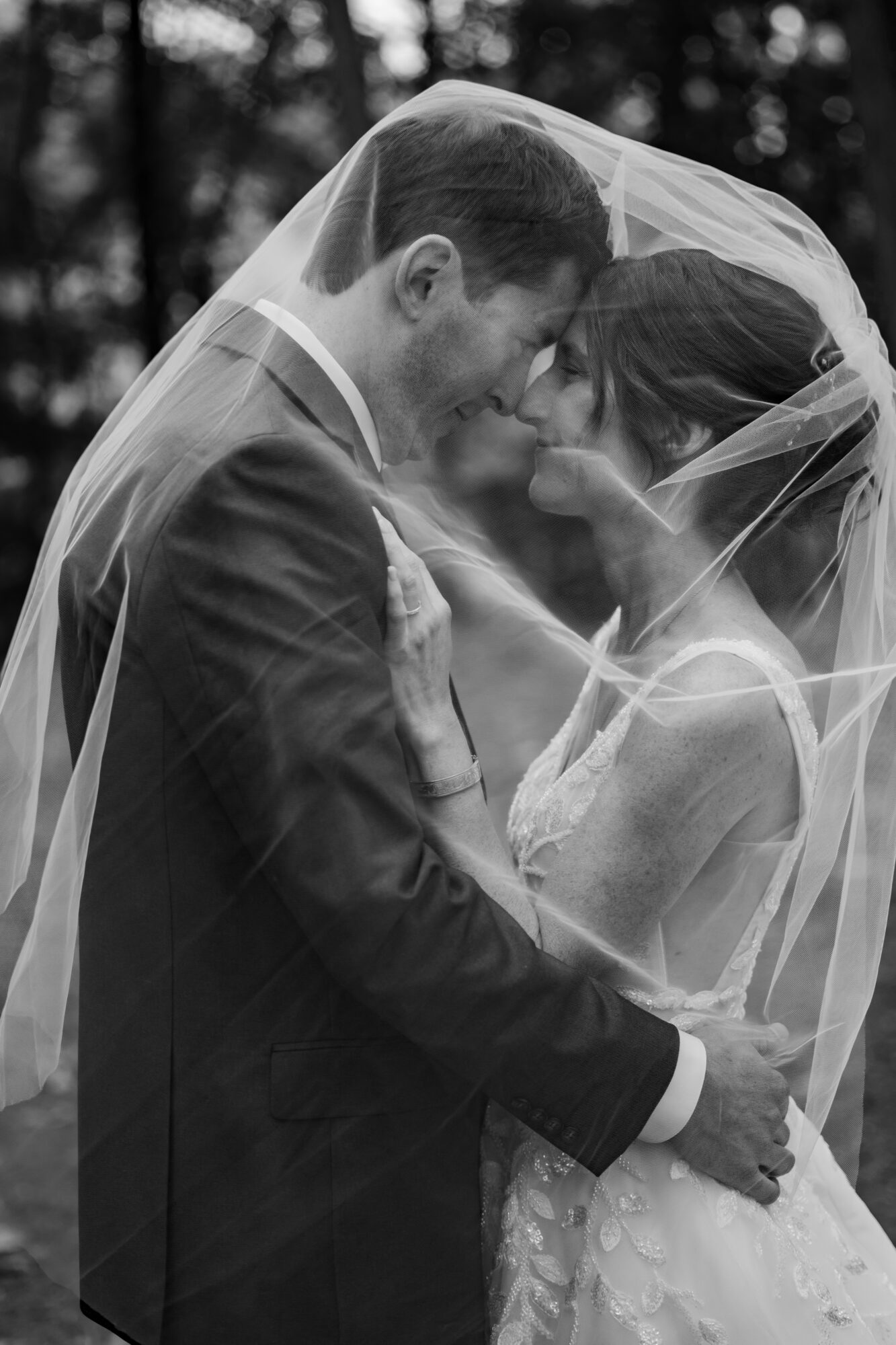A couple in wedding attire, faces close under a veil, outdoors with blurred trees in background.