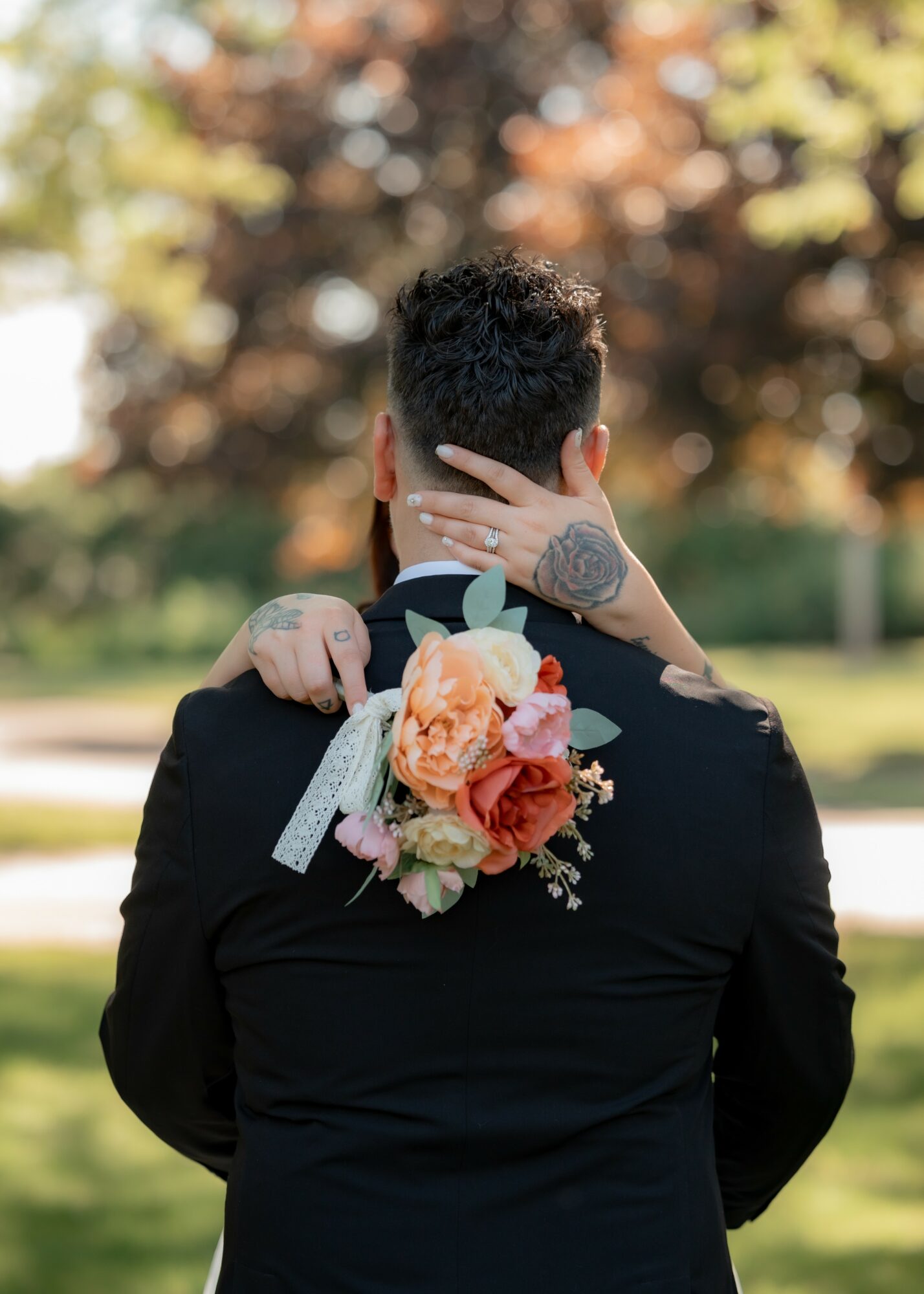 Person with short dark hair seen from behind, outdoors with trees, wearing a black jacket, holding a floral bouquet behind their neck.