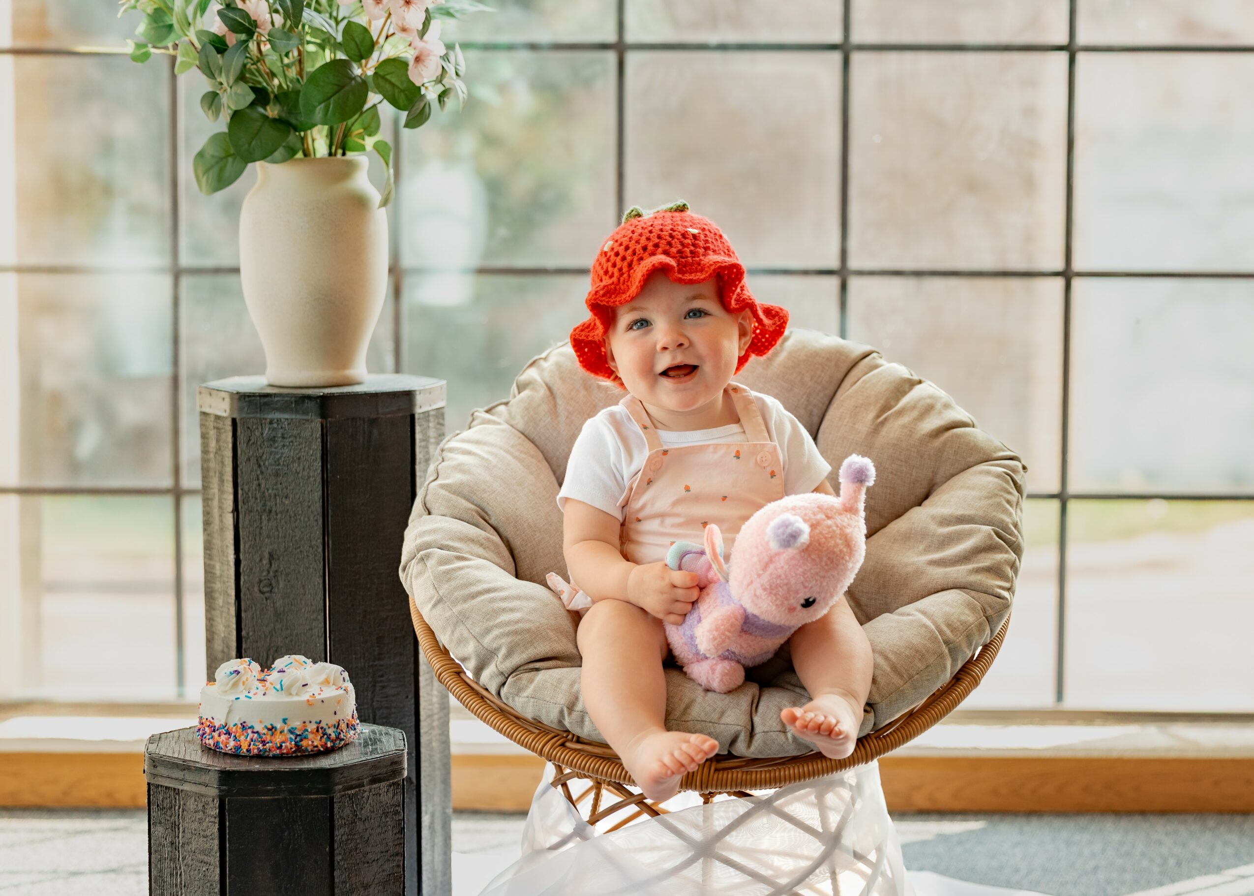 Child with red hat sitting in a round chair holding a plush toy, near a window with a vase of flowers.