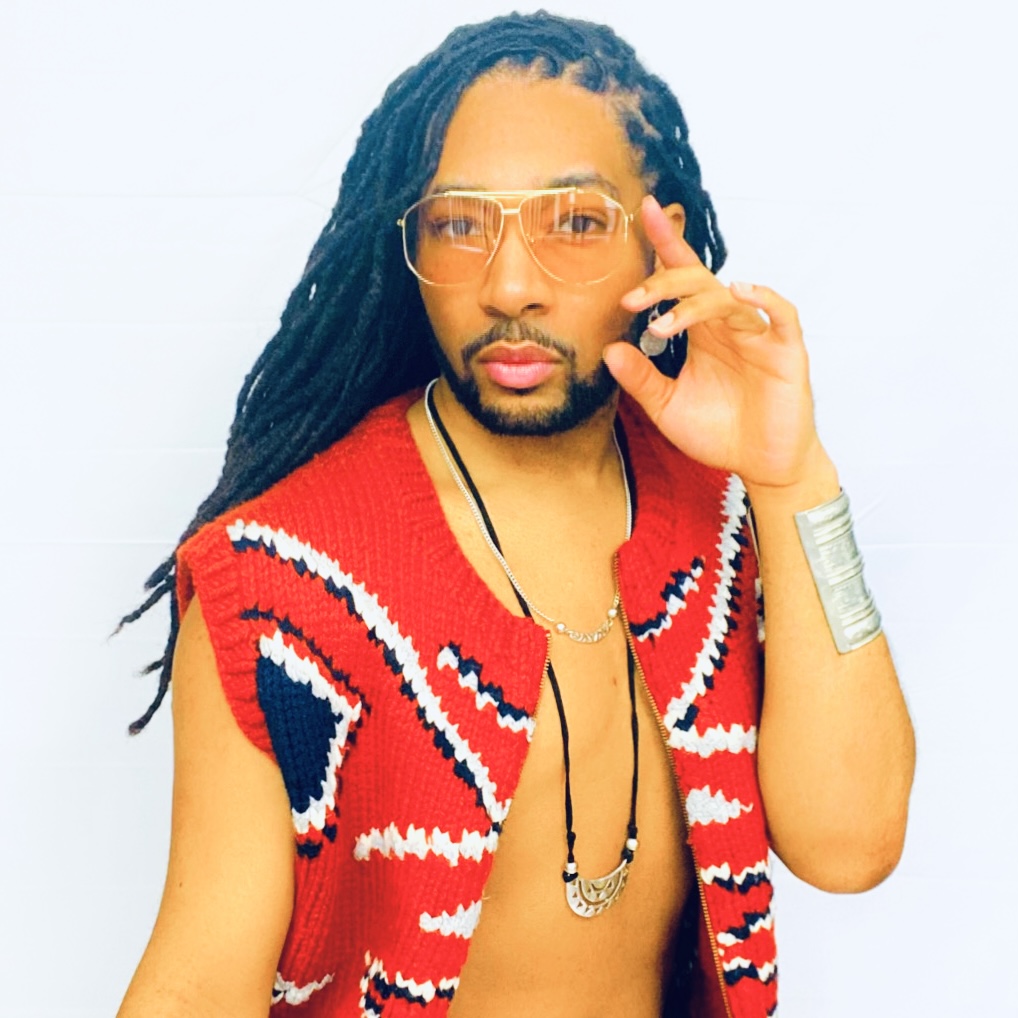 Person with long dreadlocks wearing orange-tinted glasses, a red patterned vest, and jewelry, holding a phone to their ear.