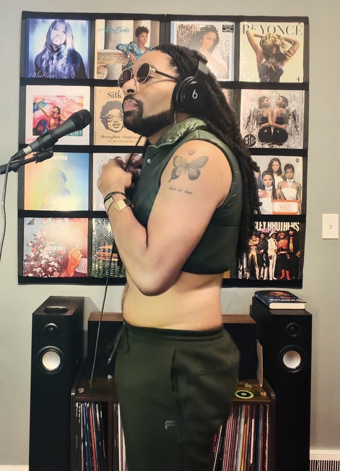 Person with long hair, sunglasses, headphones, and a tattoo on arm, standing in front of a wall with album covers, singing into a microphone.