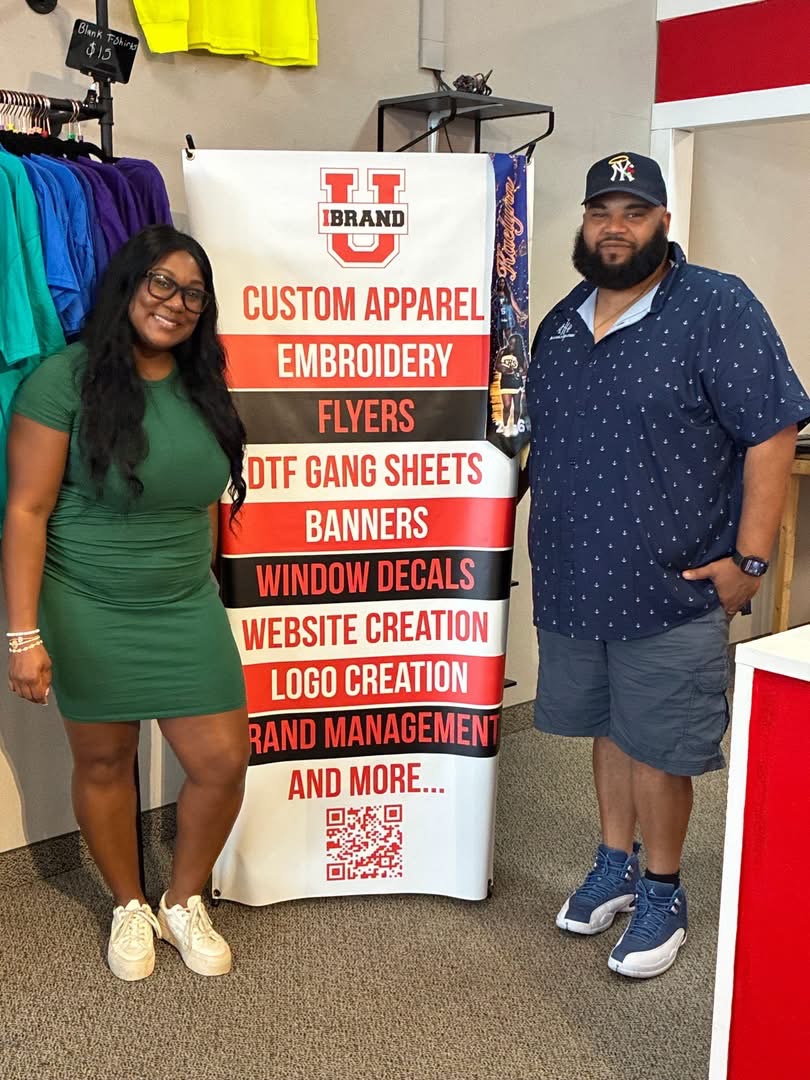 Two people standing next to a vertical sign with various promotional services listed, inside a store with colorful clothing in the background.