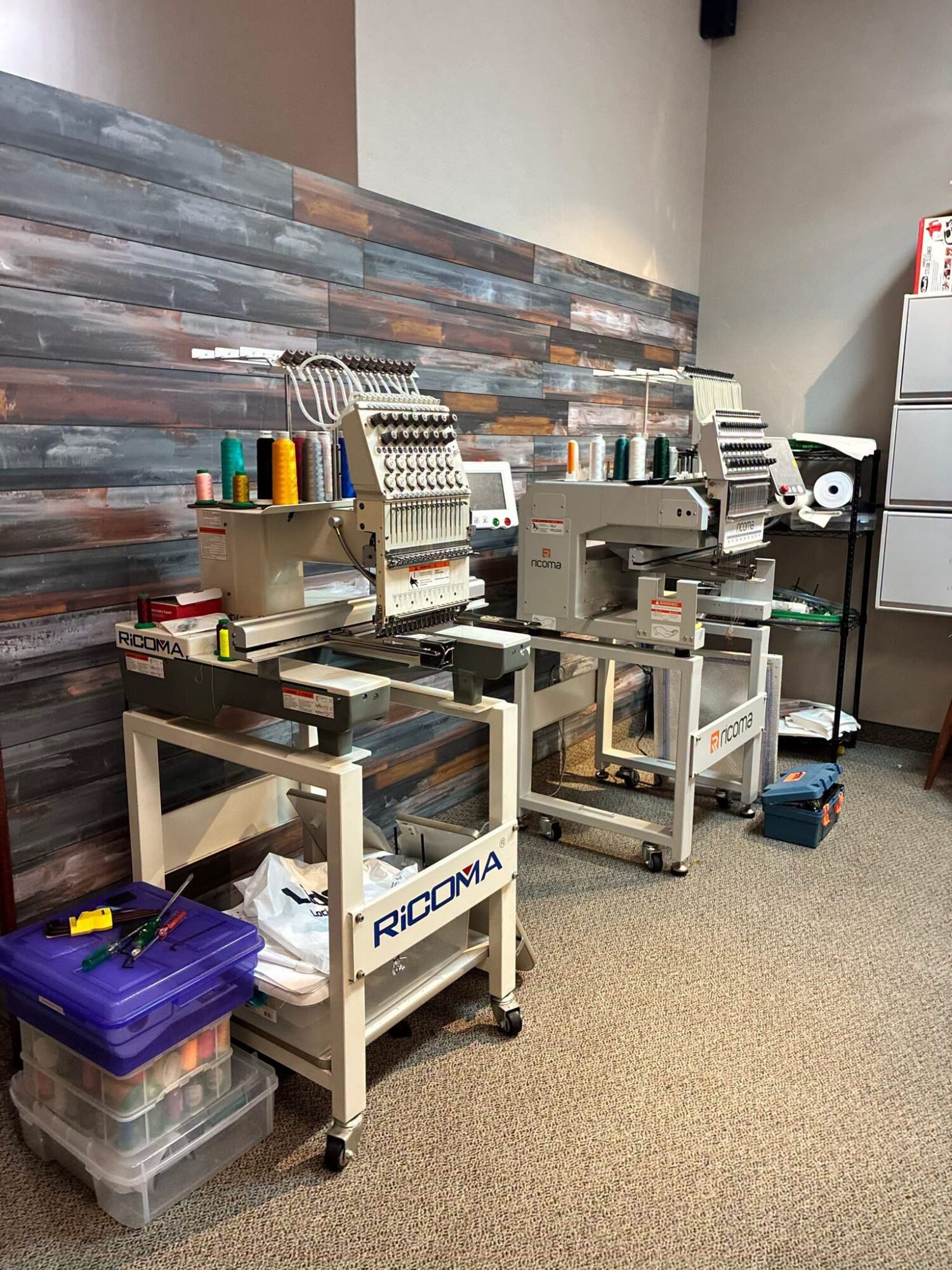 Three embroidery machines on carts in a room with a wood-paneled wall and carpeted floor.