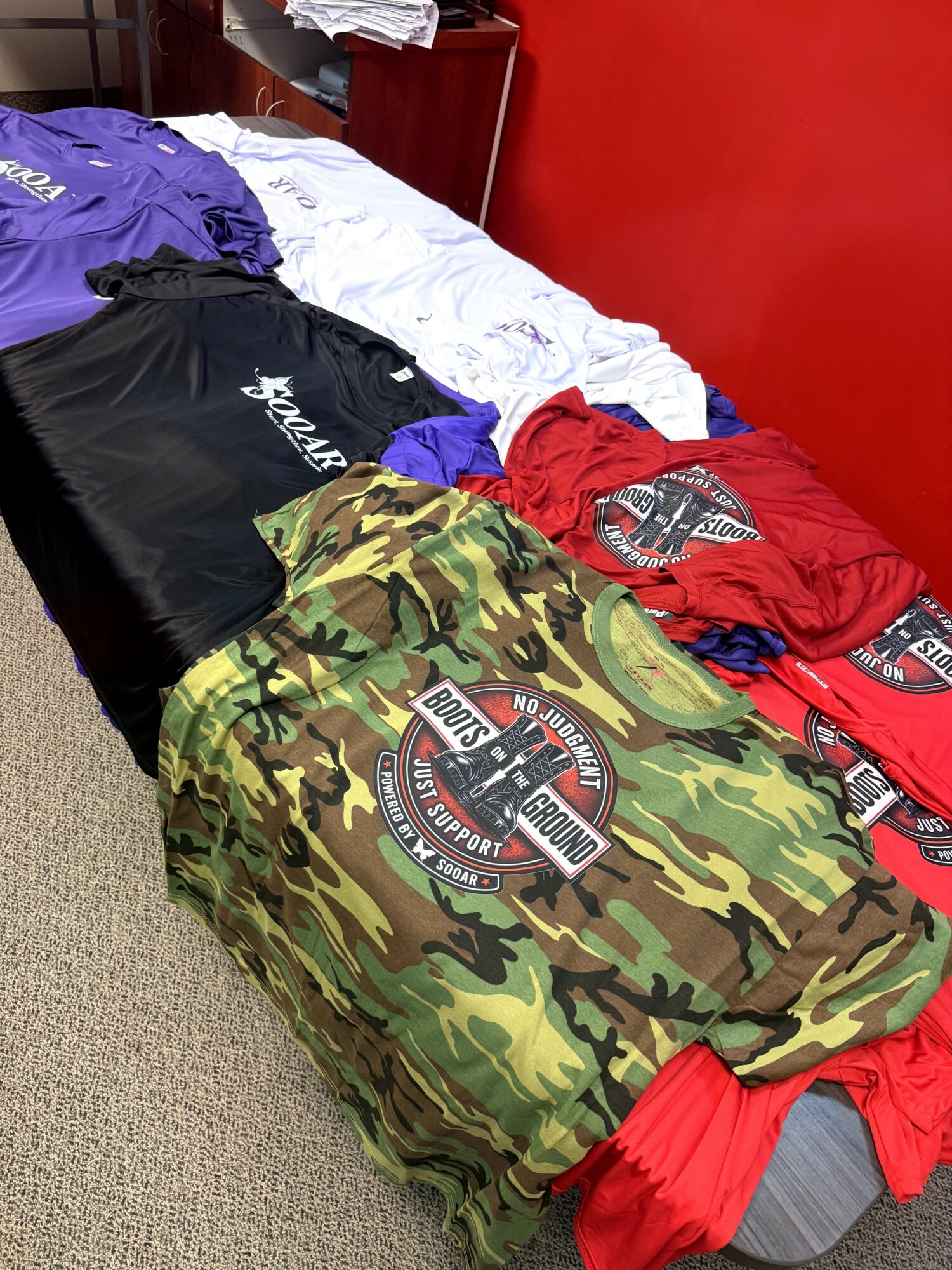 Clothes and a camouflage bag with a logo on a bed with white sheets and a red wall behind.