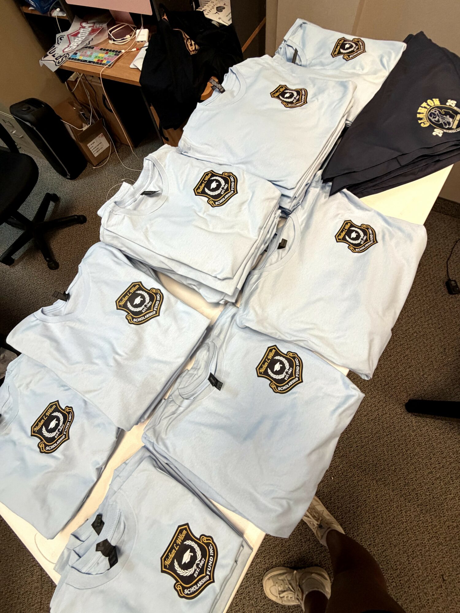 Table with folded white and black clothing items, each with a camera logo patch, in an office setting.