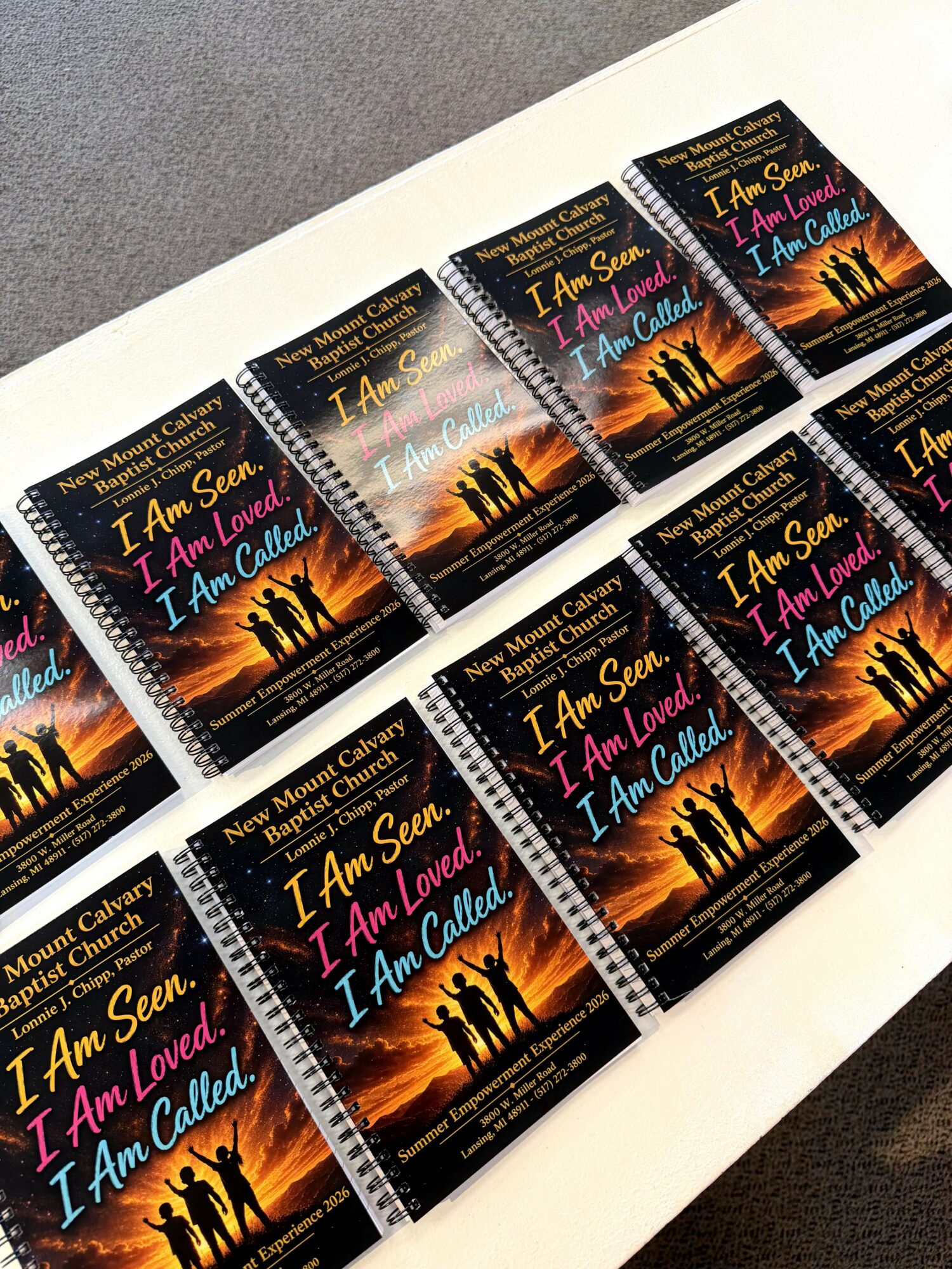 Multiple copies of a book titled 'I Am Seen, I Am Loved, I Am Called' arranged on a white surface, showing a sunset with silhouettes of people and trees.