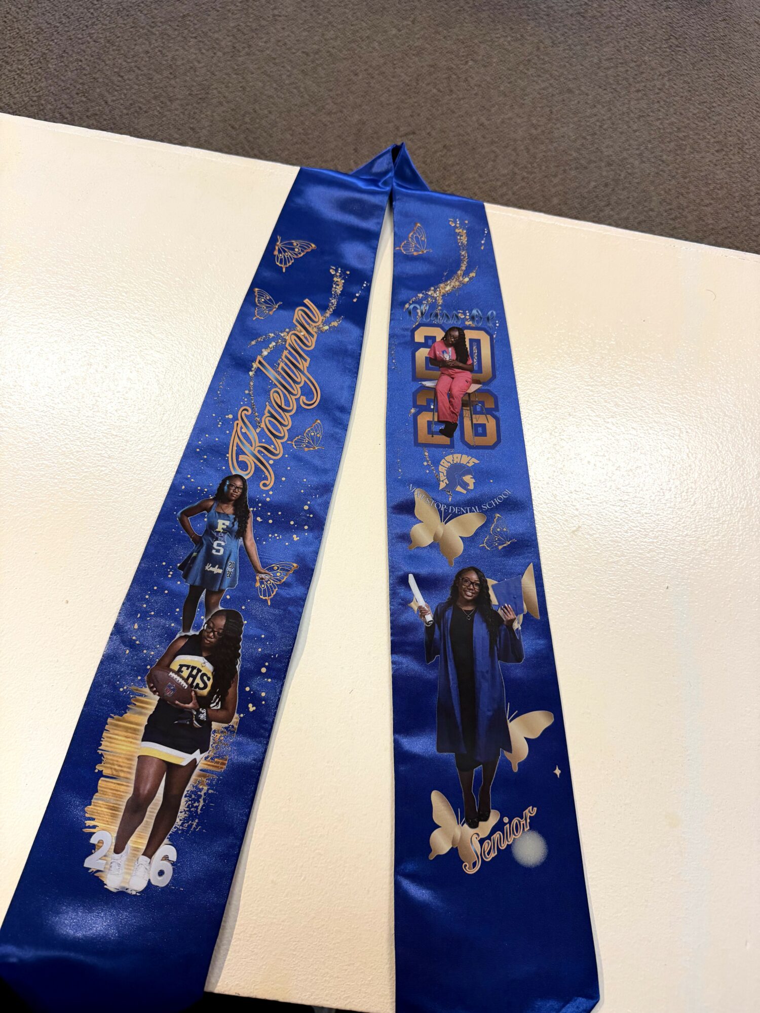 Two blue graduation sashes with images of students, butterflies, and decorative text, laid on a white surface.