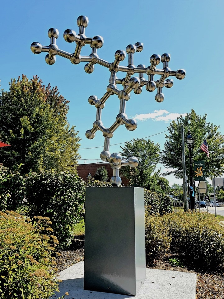 Metal sculpture resembling a molecular structure on a pedestal outdoors, with trees and a blue sky in the background.