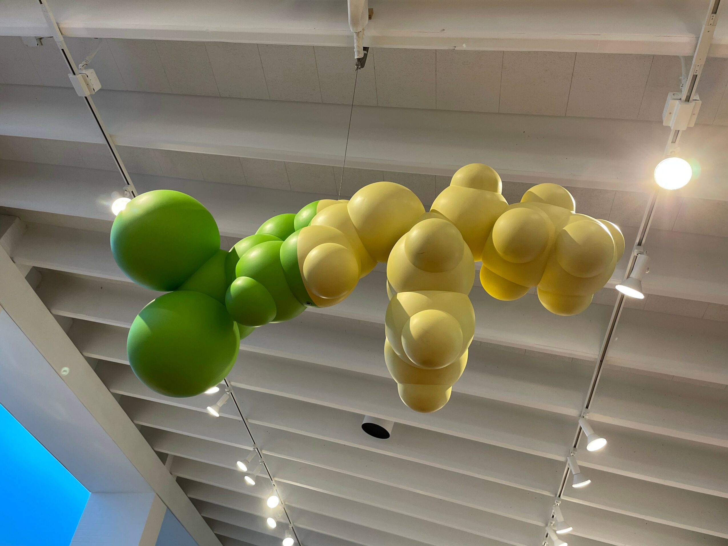 Cluster of yellow and green spherical objects hanging from a ceiling with white beams and lights