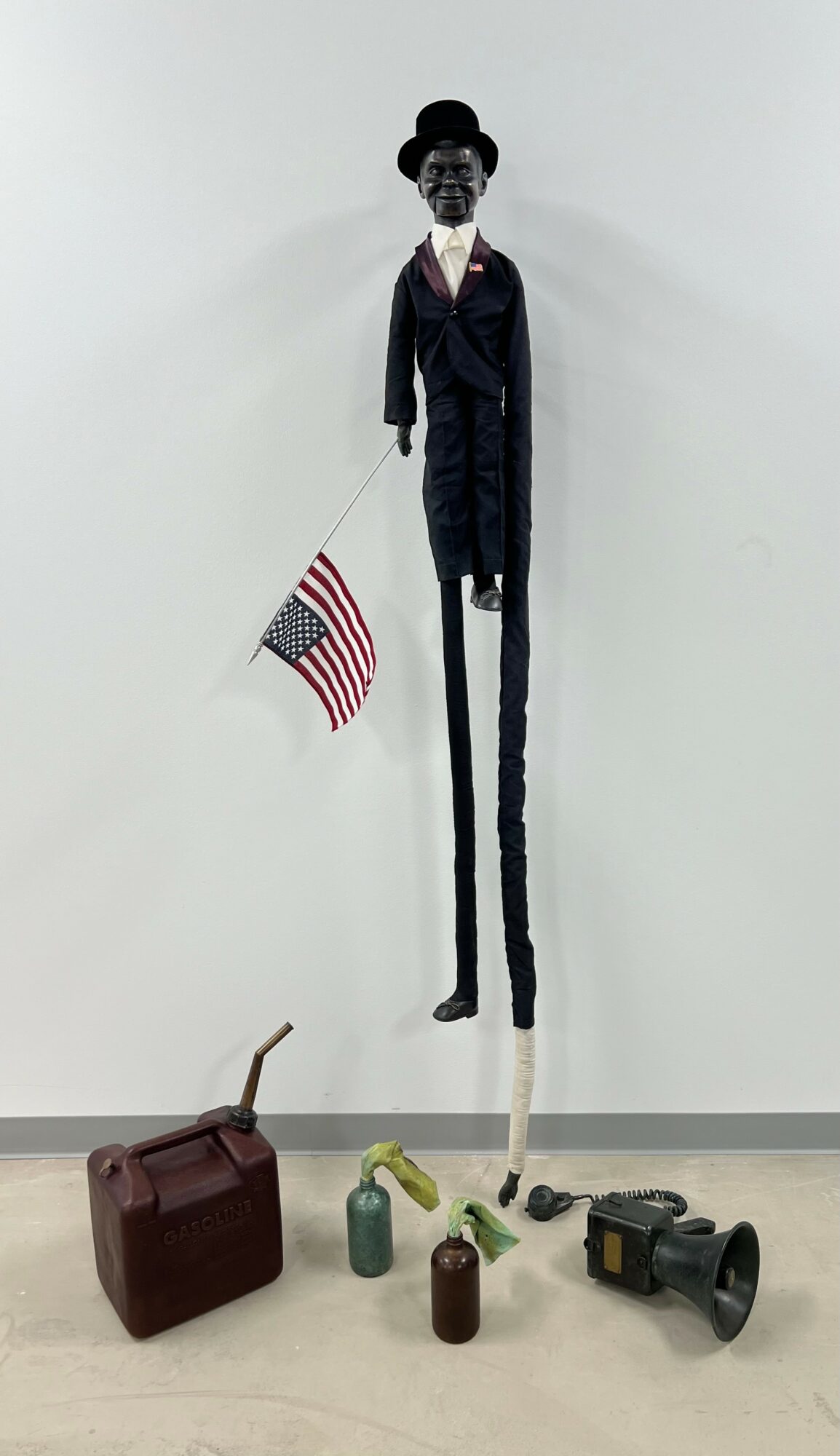 Tall puppet with a top hat, suit, and American flag, surrounded by a briefcase, bottles, and a megaphone, against a plain wall.