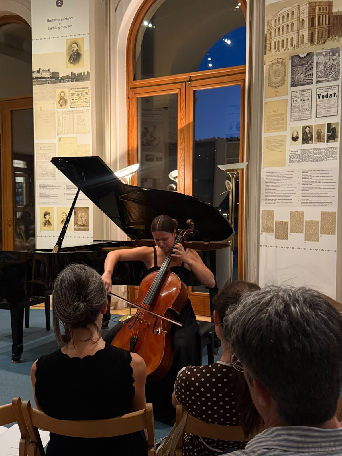 Person playing cello in a room with large windows and informational posters on the walls, audience seated nearby.