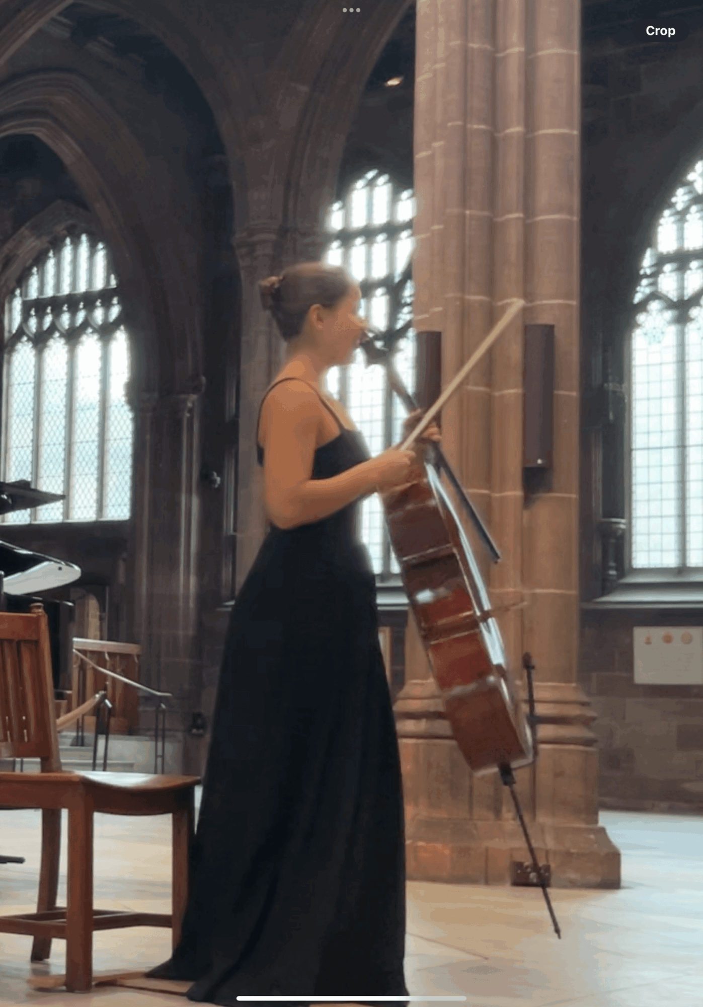 A woman in a black dress stands inside a large, historic building with tall windows and stone columns, holding a cello and bow.