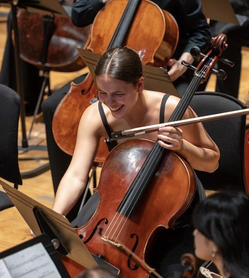 Woman playing a cello, smiling, with other musicians and instruments in the background.
