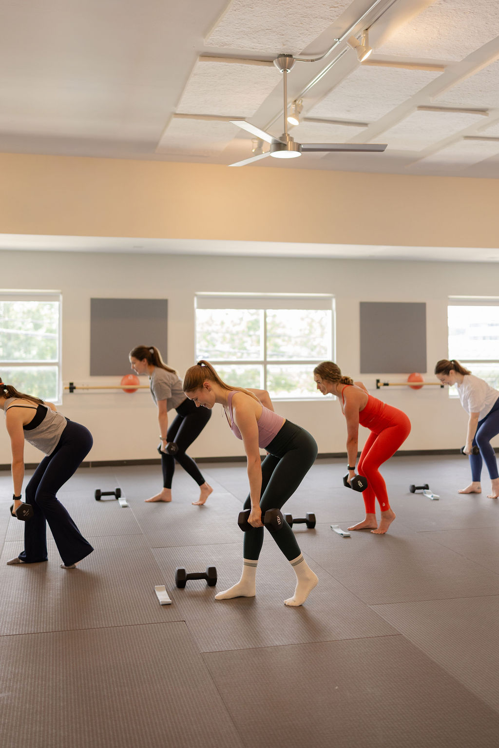 Group of women exercising with dumbbells in a bright indoor studio, stretching and bending forward, windows in background.
