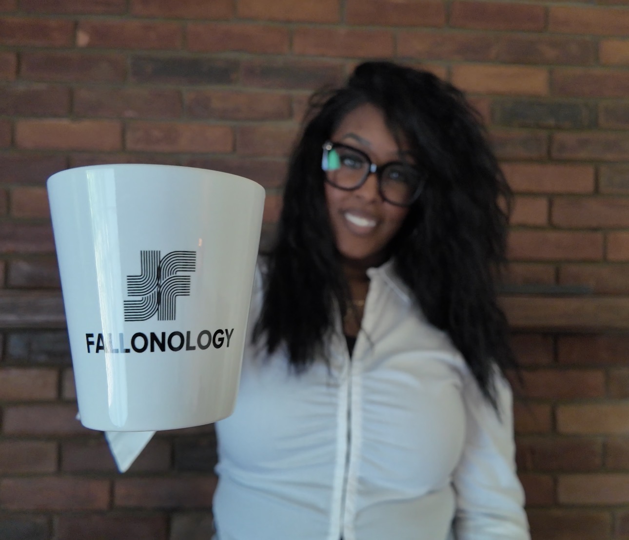 Woman with glasses holding a white mug with a logo, standing in front of a brick wall.