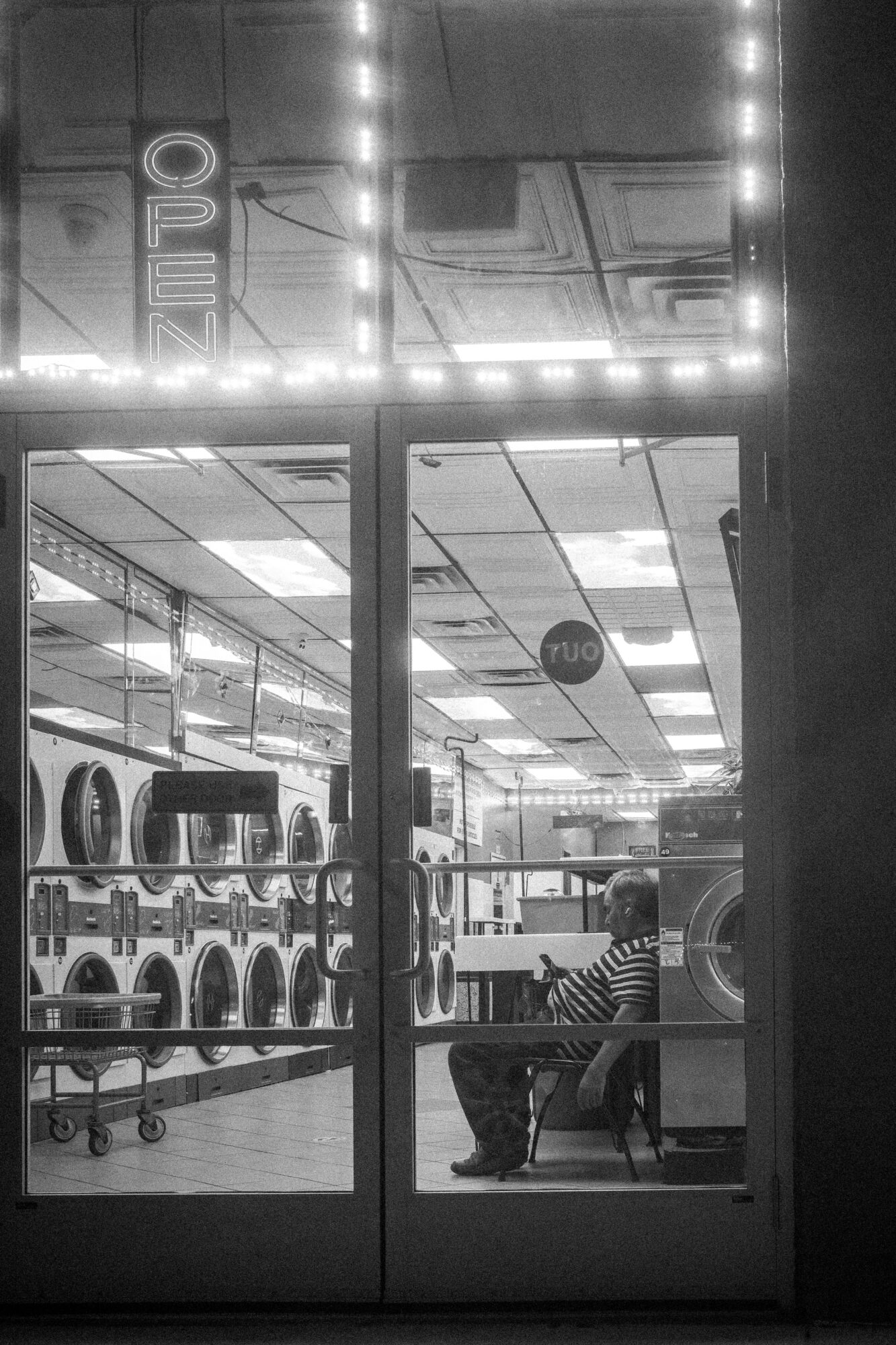 Person sitting on a bench inside a store, viewed through glass doors, with washing machines visible inside, black and white.