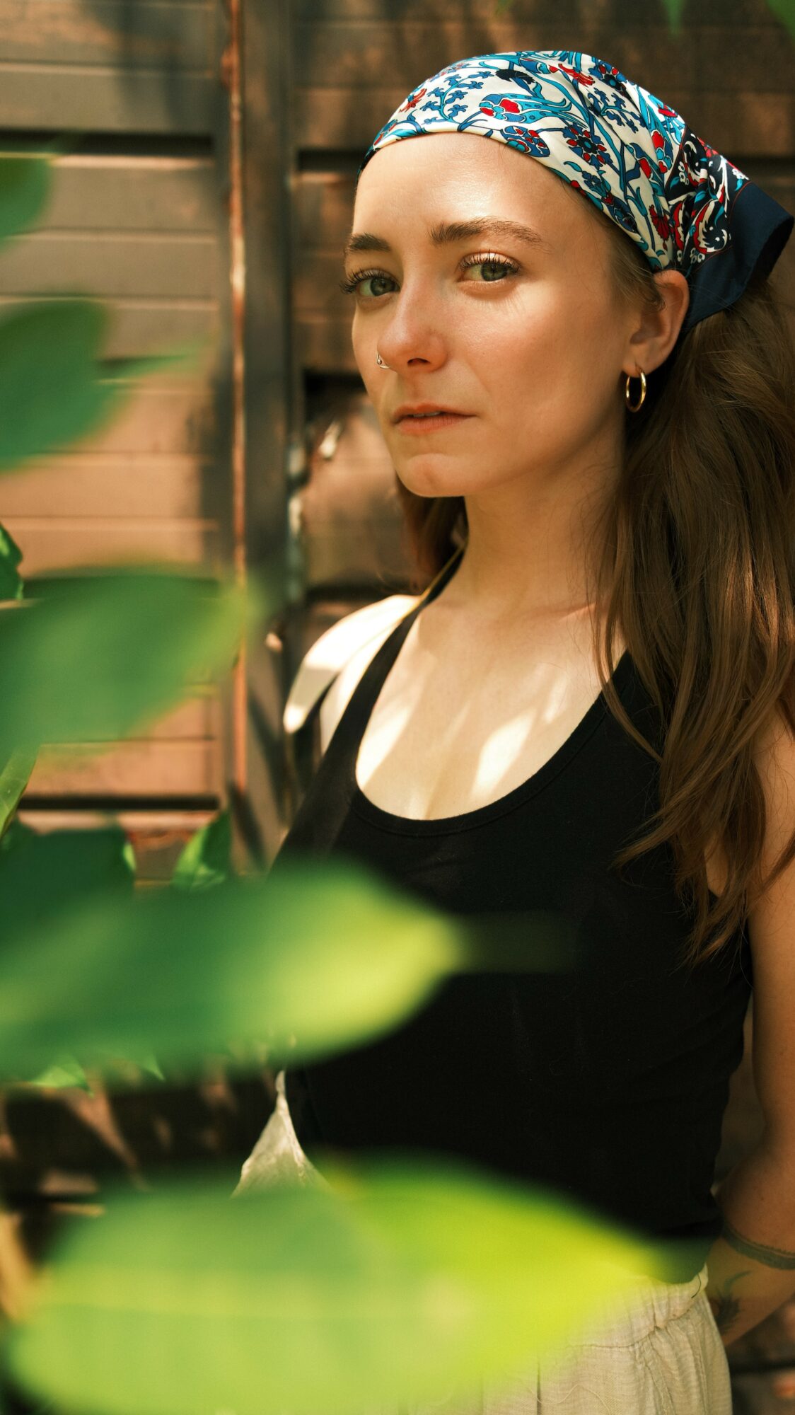 Woman with a colorful headscarf and hoop earrings looks to the side outdoors, surrounded by green leaves.
