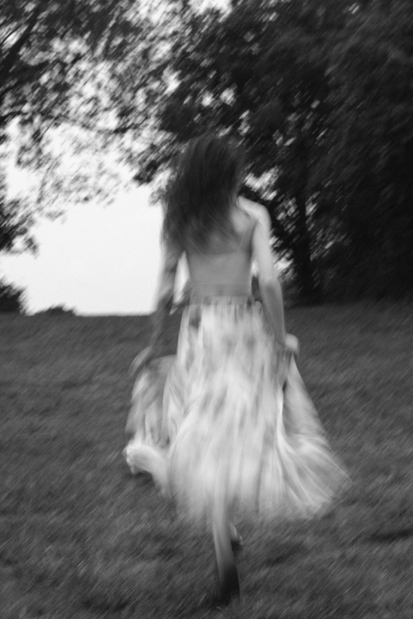 Person with long hair walking away on a path surrounded by trees, in black and white.