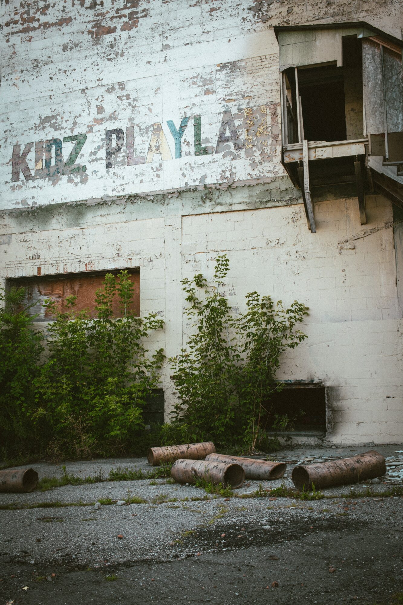 Old building with a sign reading 'KIDZ PLAYLAND' partially visible, surrounded by overgrown bushes and debris on the ground.