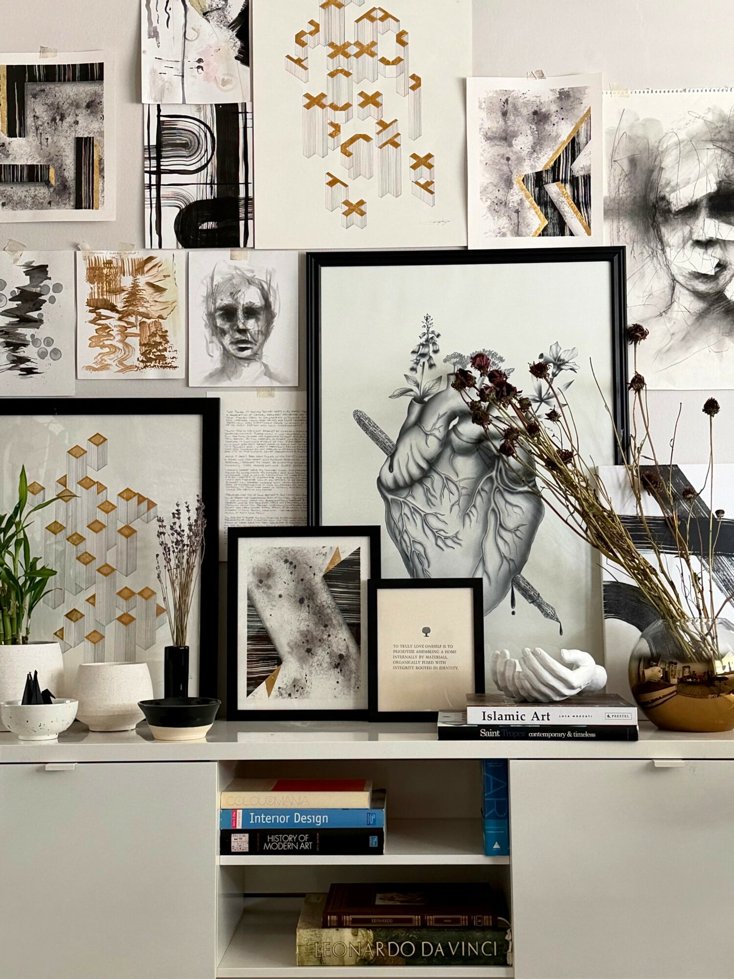 Wall decorated with various art prints, framed drawings, and photographs, with a white shelf holding books, vases, and dried flowers.