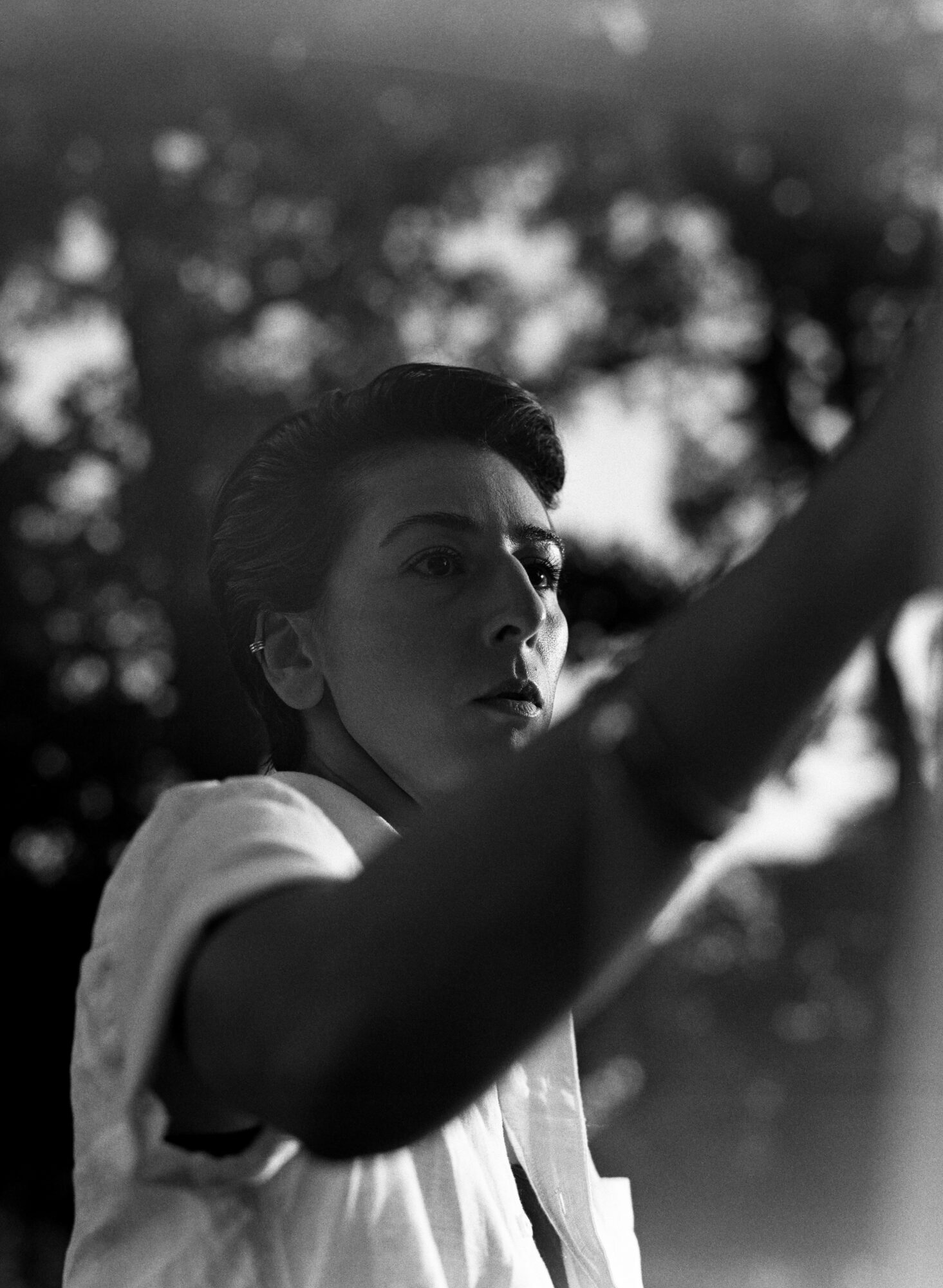 A woman with short hair looks to the side outdoors, with trees and sky in the background.