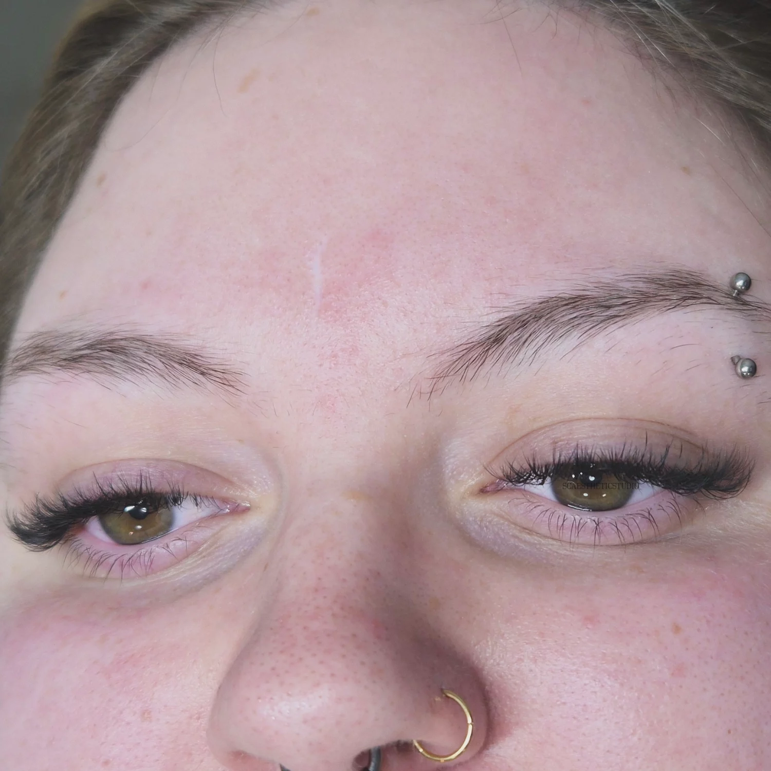 Close-up of a person's face with eyes open, showing eyebrows, eyelashes, and a nose piercing.