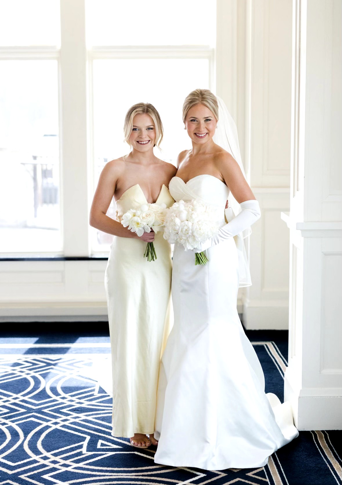 Two women in wedding dresses holding bouquets, standing indoors near large windows, smiling, with a patterned carpet.