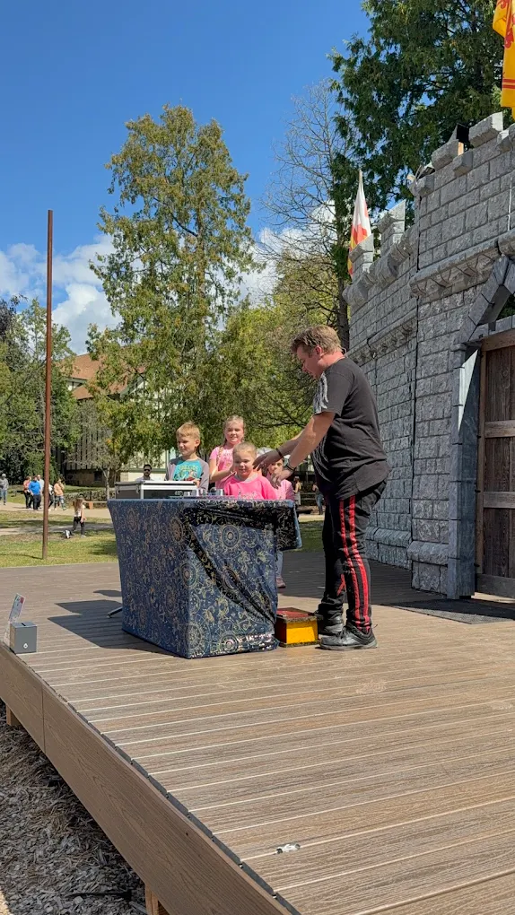 Person standing next to a table with children seated, outdoors on a wooden platform, with trees and a stone wall in background.