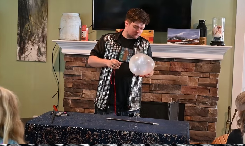 Young person performing a science experiment with a large glass container in a room with a fireplace.