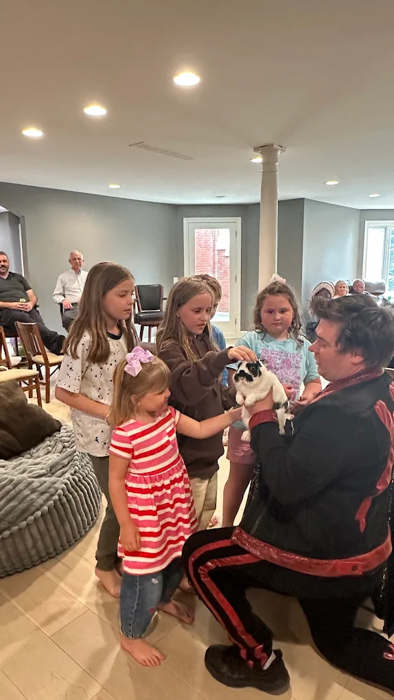 Group of children gathered around a person holding a small animal indoors, with windows and a column in the background.