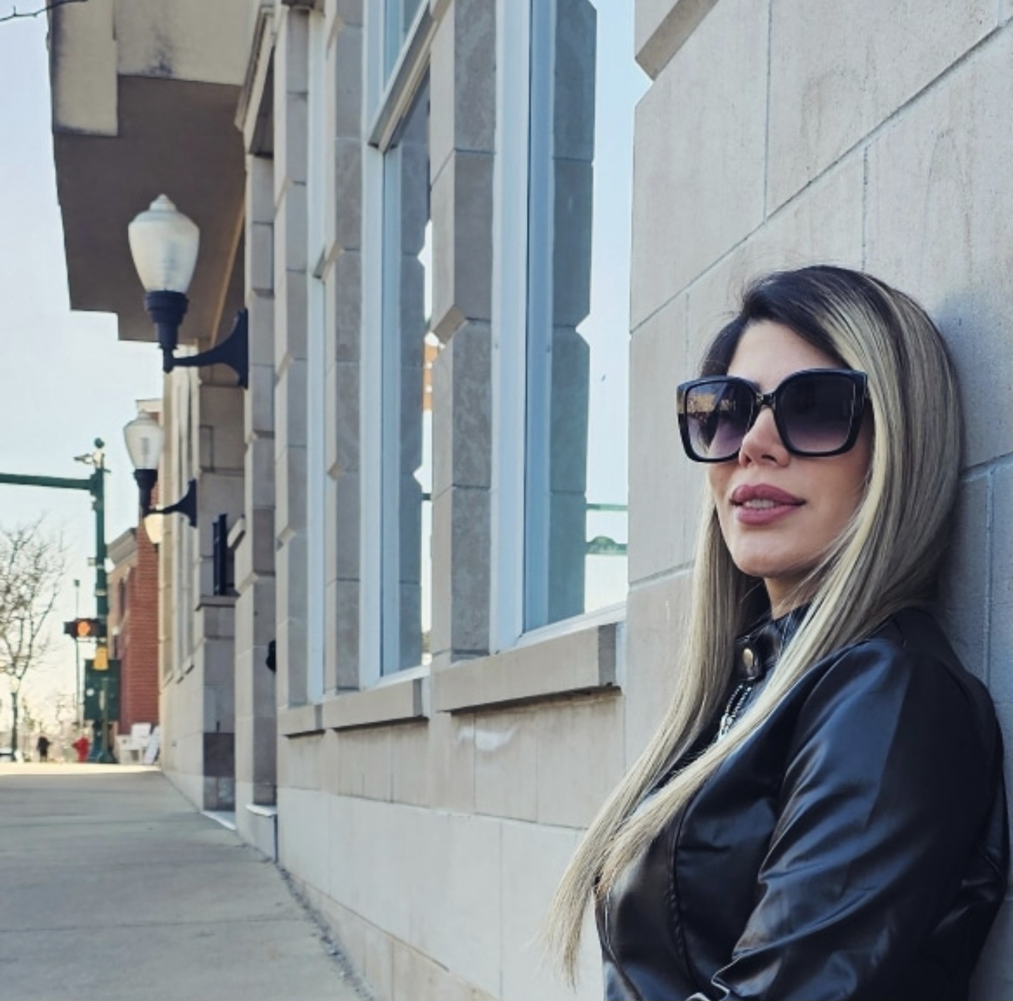 Woman with long hair and sunglasses leaning against a wall outside, wearing a black jacket.