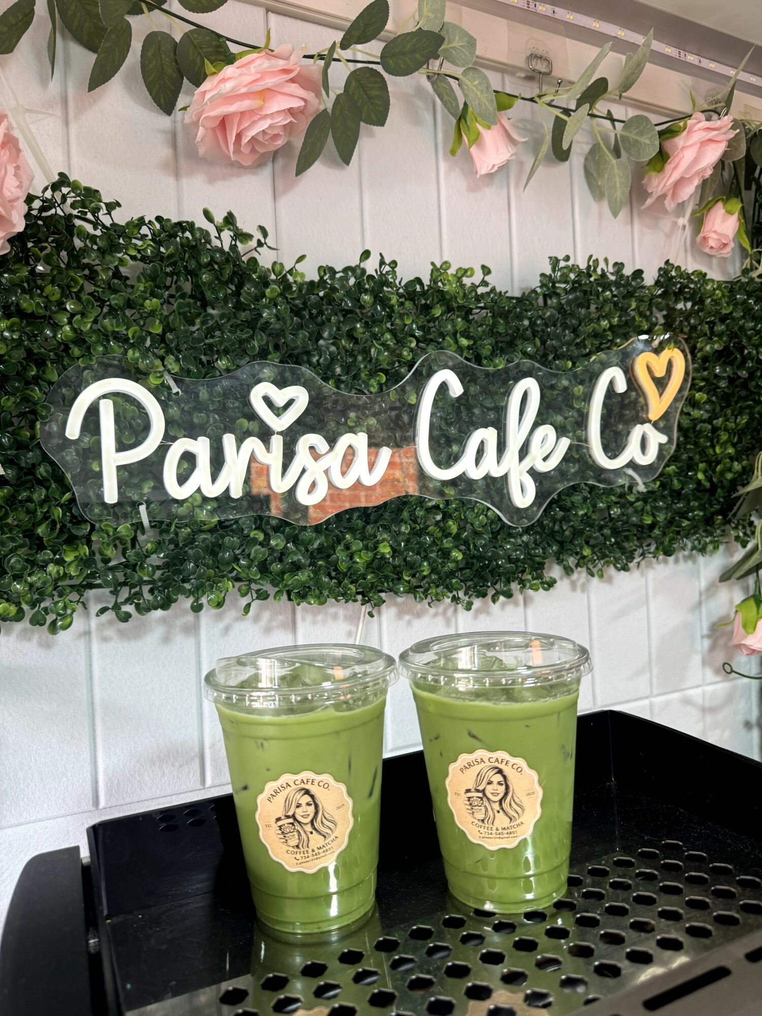 Sign reading 'Parisa Cafe' on a green leafy background with pink flowers above, two green drinks in front, on a black tray.