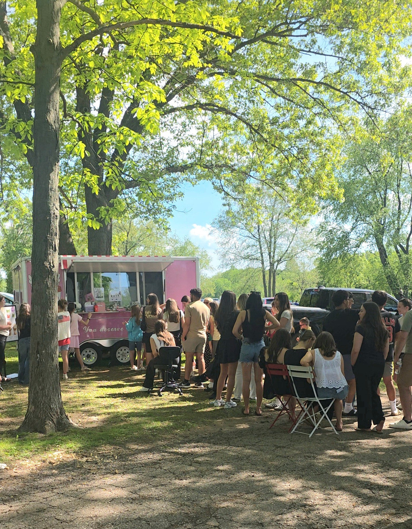 Group of people gathered outdoors near a pink food truck, with trees and sunlight in the background.
