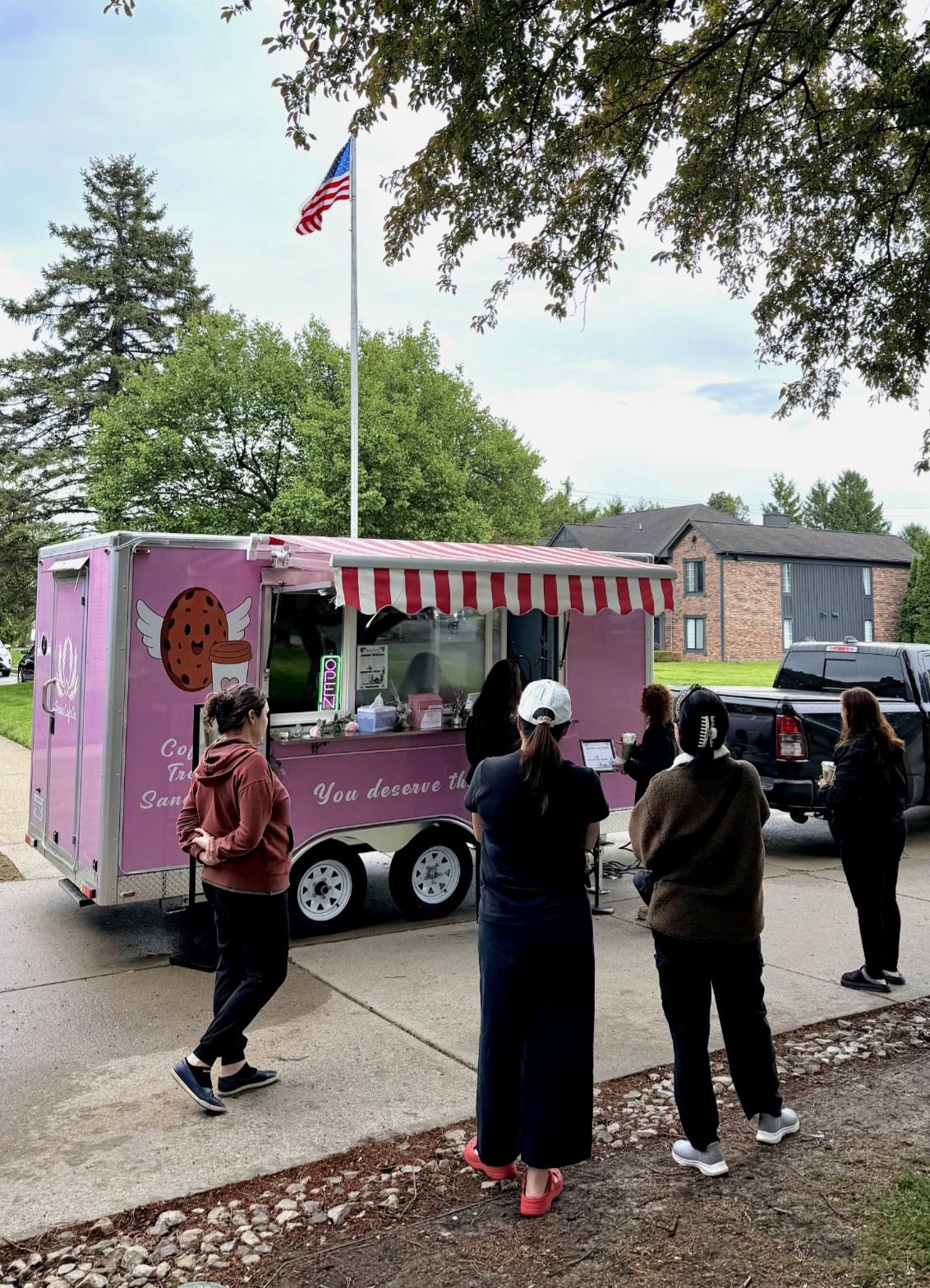 People line up at a pink ice cream truck with a red and white striped awning, parked on a sidewalk under trees.