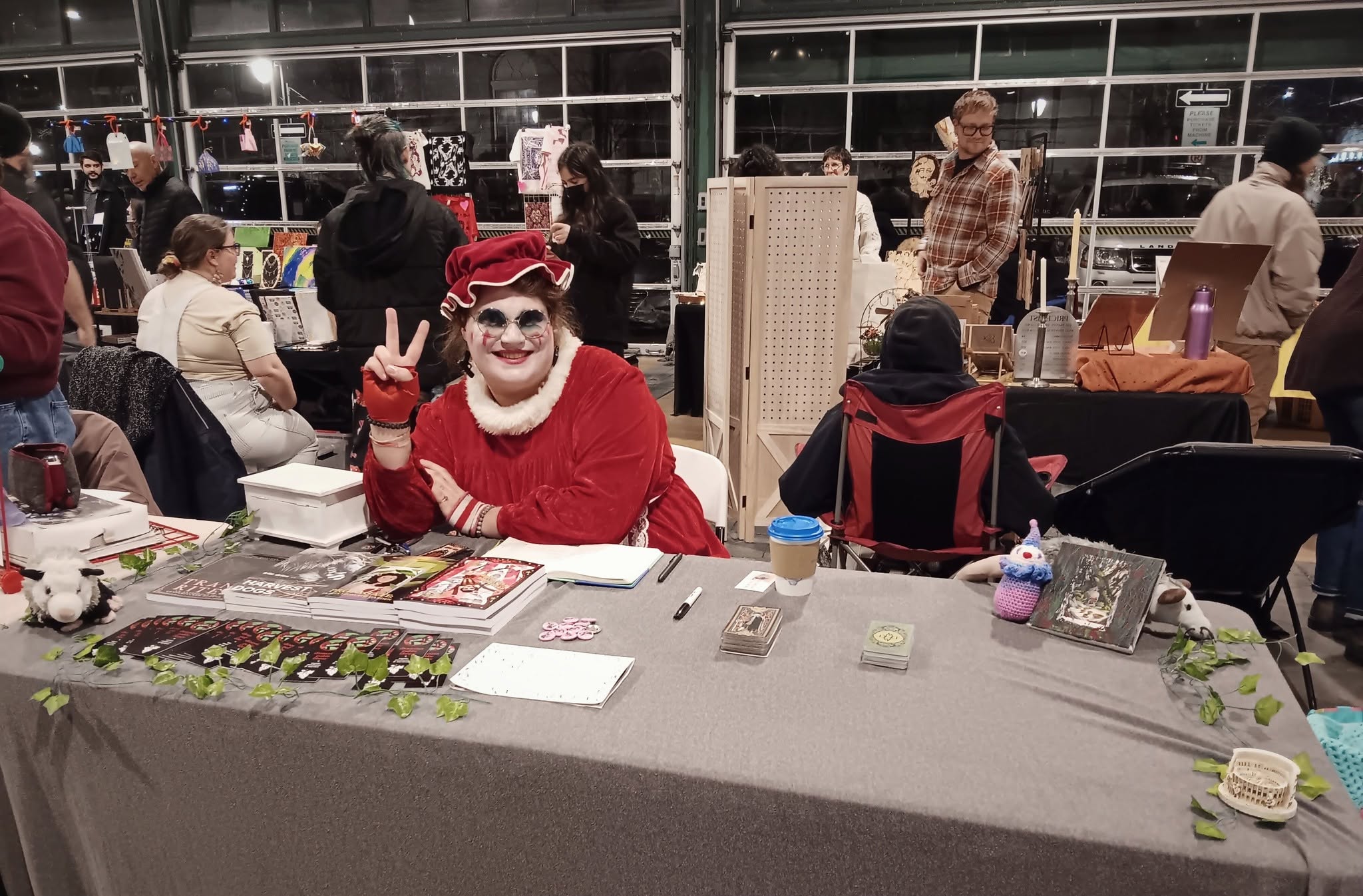Person dressed as a clown with face paint and red costume, sitting at a table in a busy indoor event space, making a peace sign.