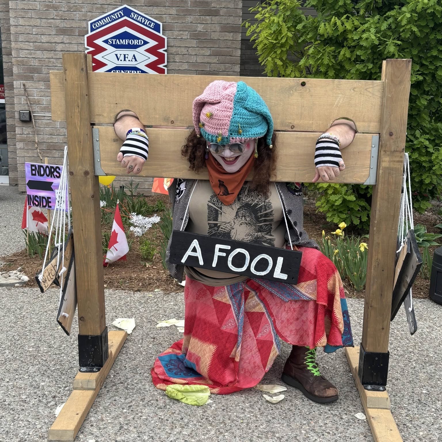 Person with colorful hat and face paint kneels behind a wooden stock, holding a sign that says 'A FOOL'.