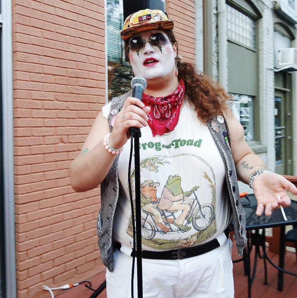 Person with makeup, sunglasses, red bandana, holding microphone, wearing graphic T-shirt, standing outside near brick wall.